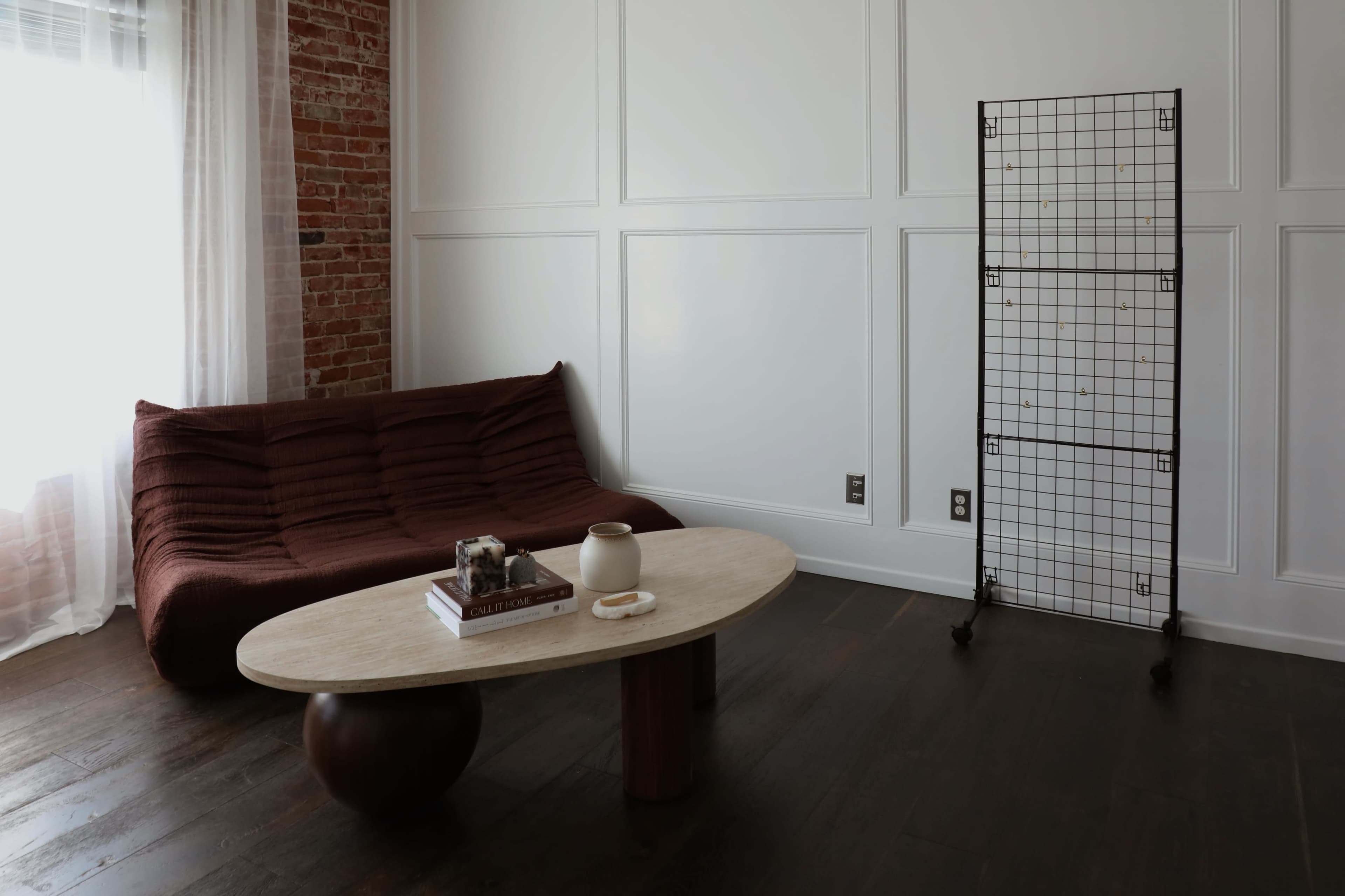 The image shows a modern living room with a maroon sofa, a round wooden coffee table, and a wire display stand against a wall with white paneling and brick accents.