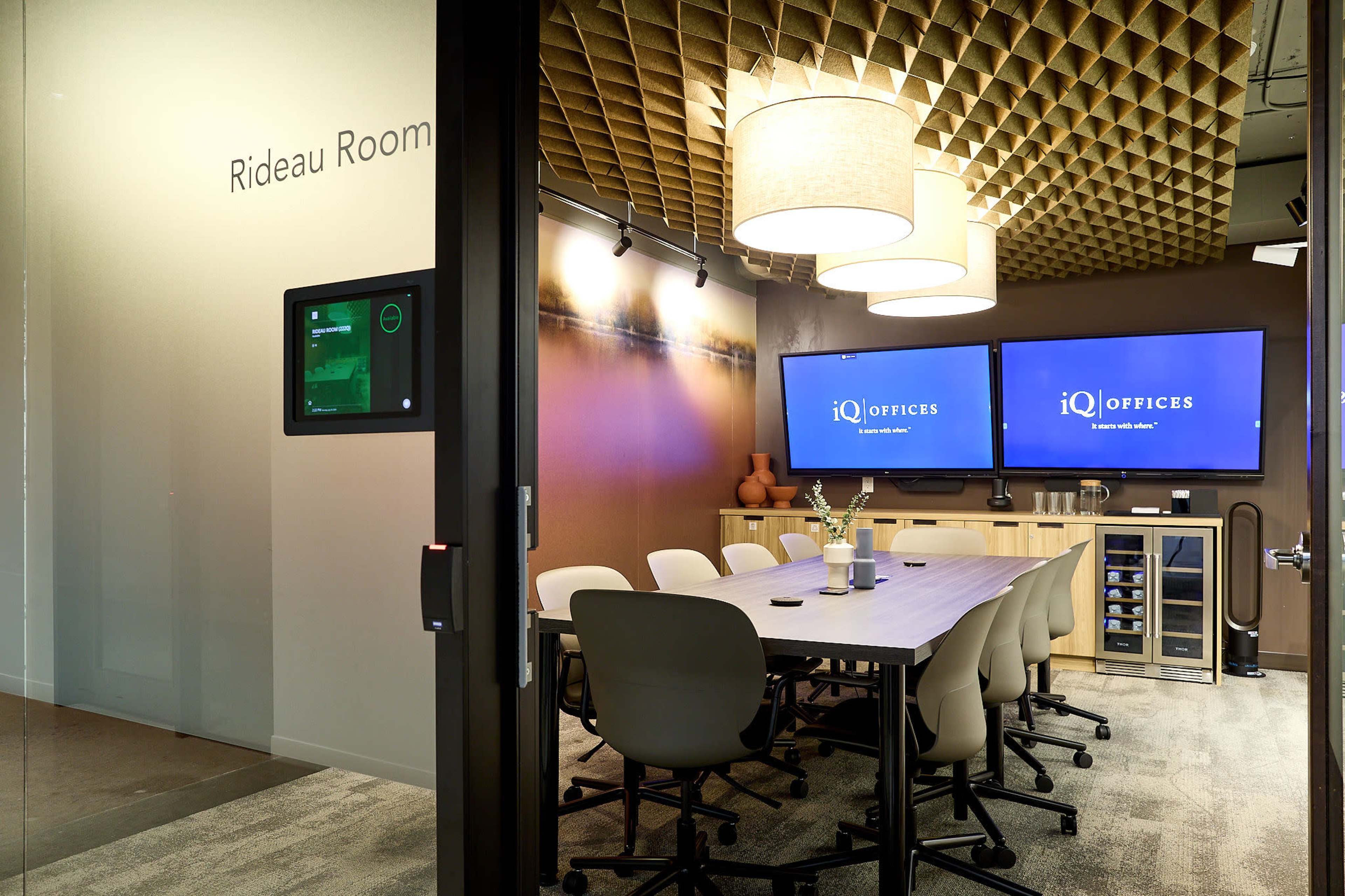 The image shows a conference room named "Rideau Room," featuring a large table, several chairs, and two wall-mounted screens displaying the "iQ Offices" logo.