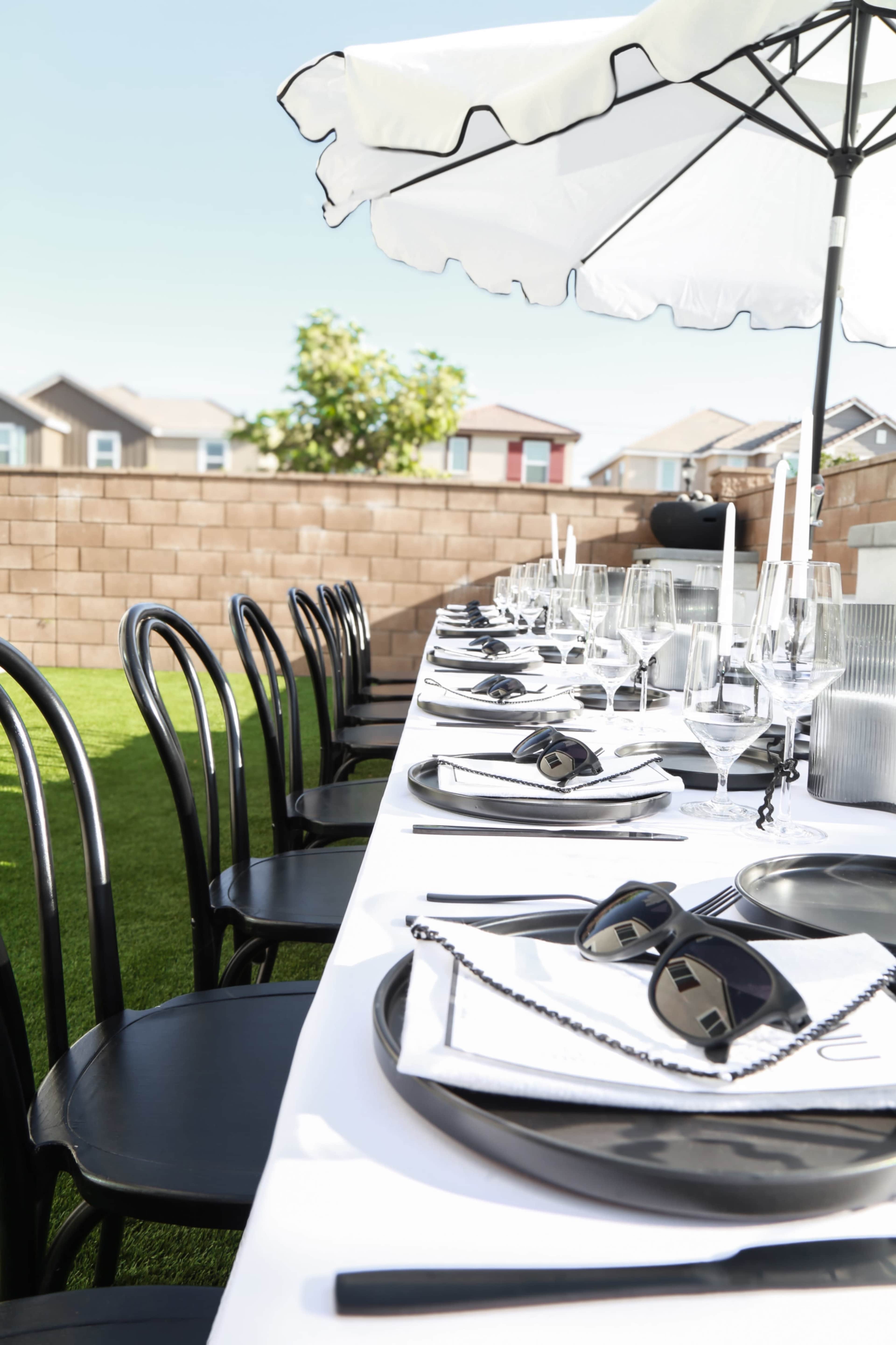 A long, elegantly set dining table with black chairs is arranged outdoors under a white umbrella in a backyard.
