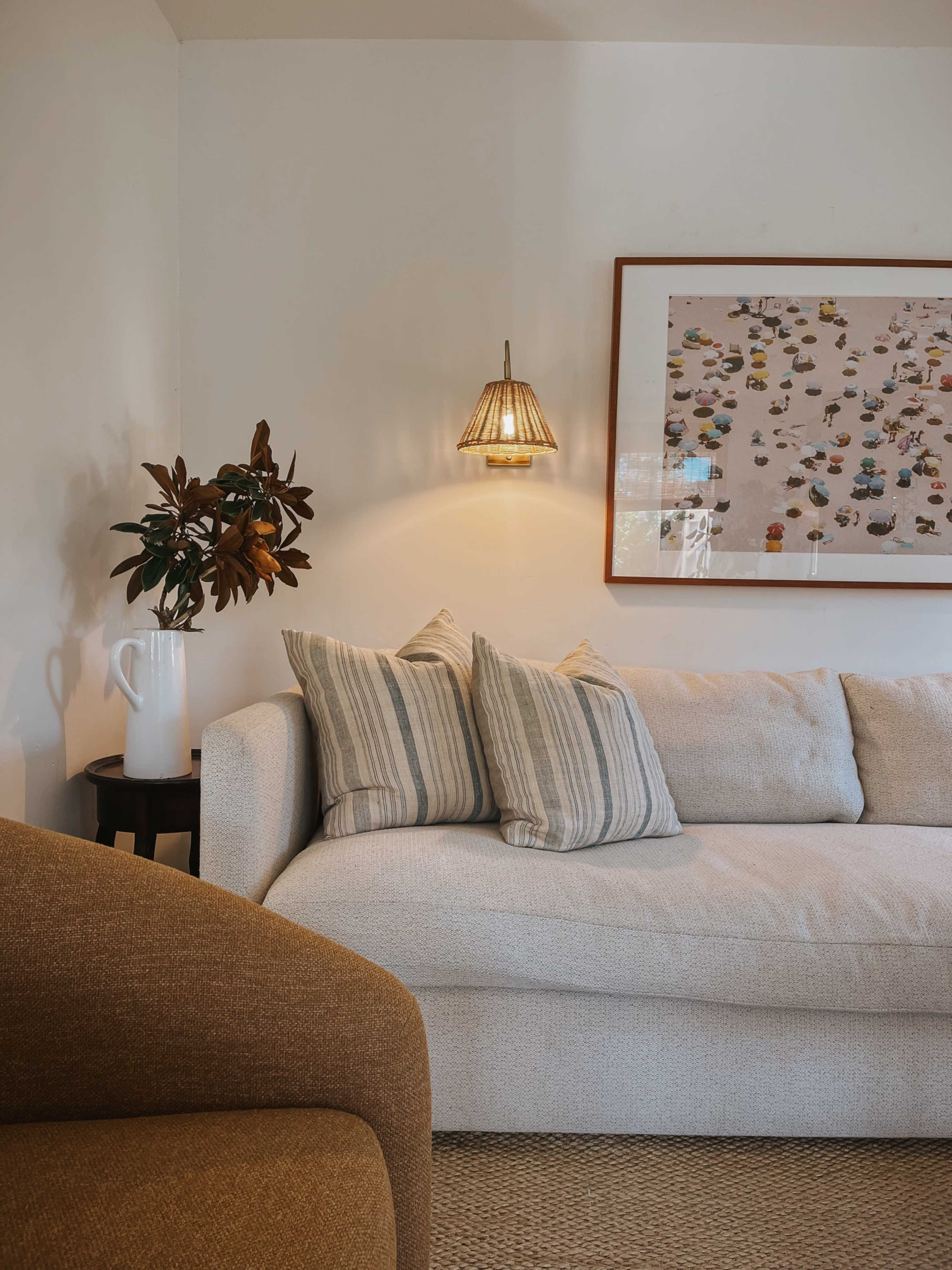 A light-colored couch with striped pillows is positioned against a wall, alongside a small table with a vase of flowers and a framed artwork depicting colorful boats.