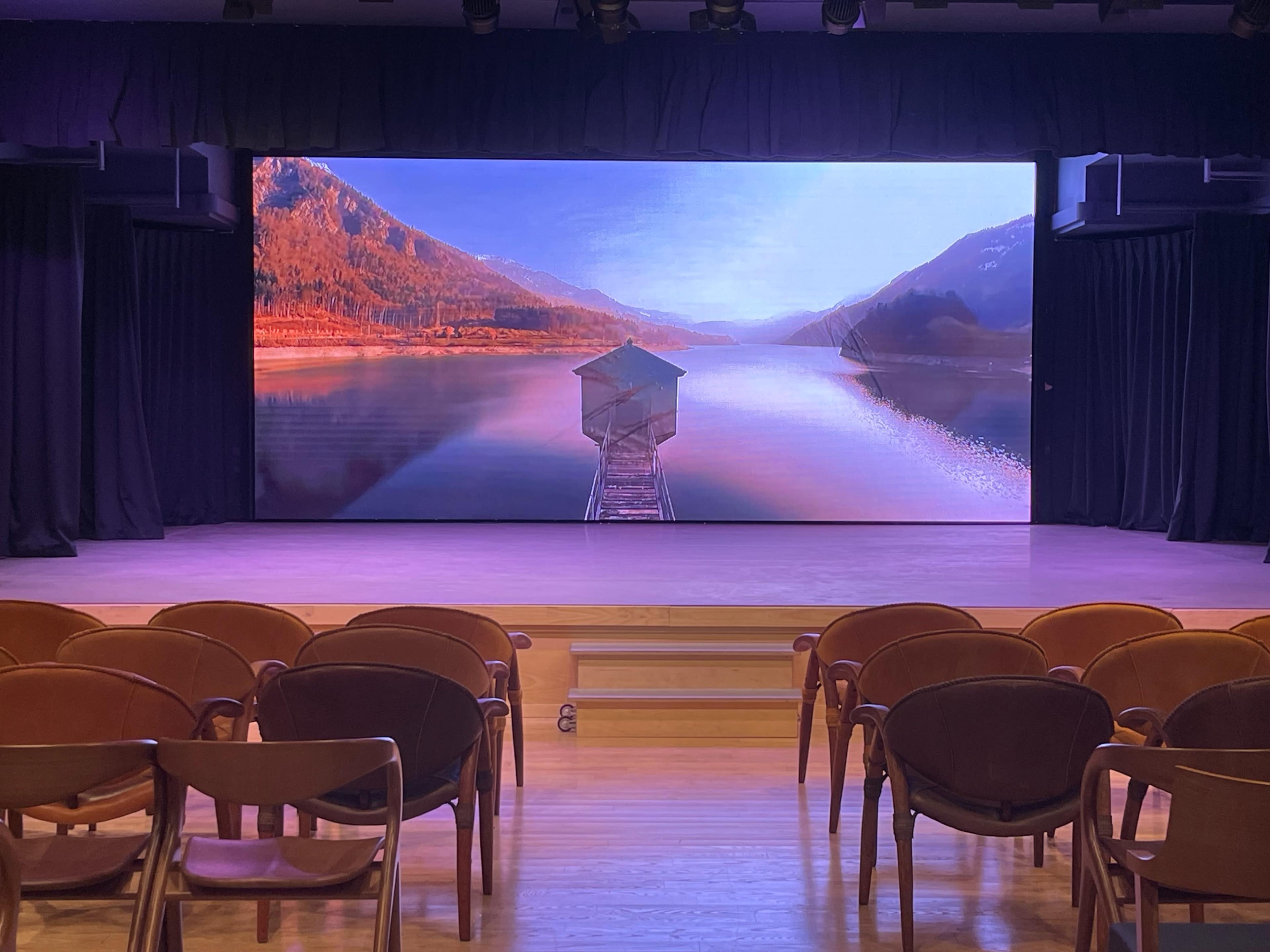 The scene features a large screen displaying a serene landscape of a lake with a wooden structure extending into the water, surrounded by mountains, in a theater setting with empty brown chairs facing the screen.