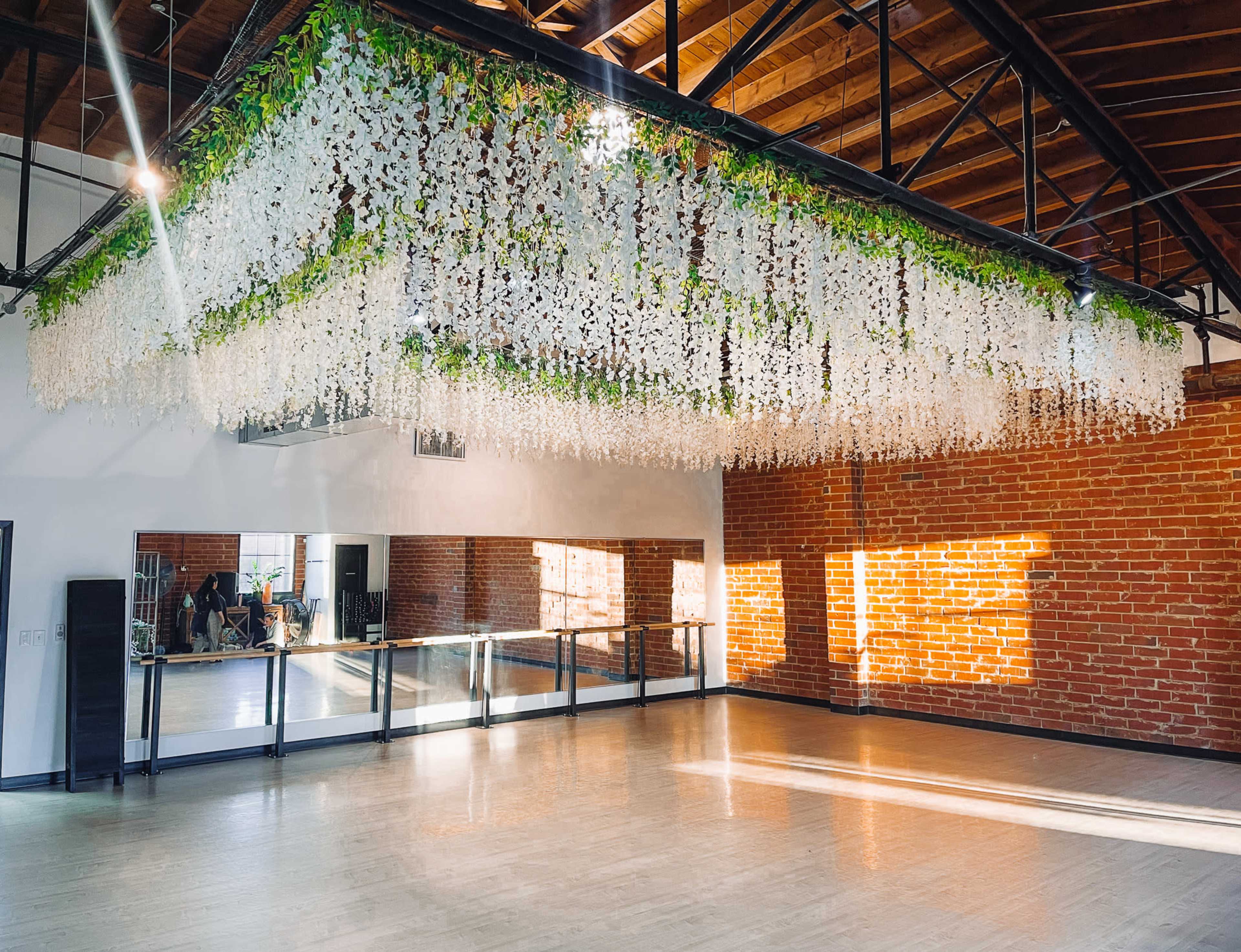 The image shows a spacious dance studio with a large floral-inspired ceiling installation and hardwood floors, featuring brick walls and mirrored panels.