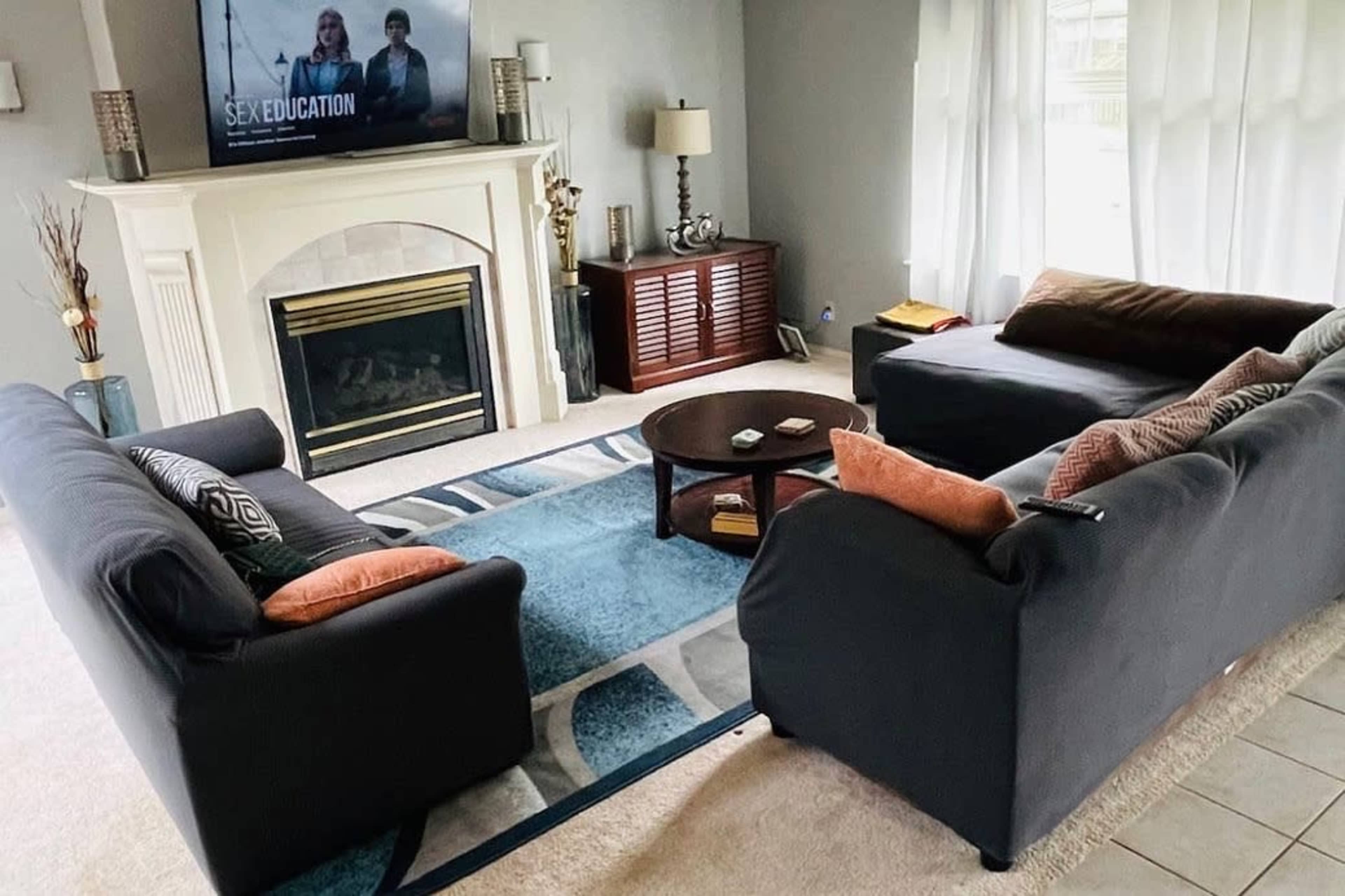 The living room features two black couches with orange pillows facing a coffee table, a fireplace, and a television displaying the show "Sex Education."