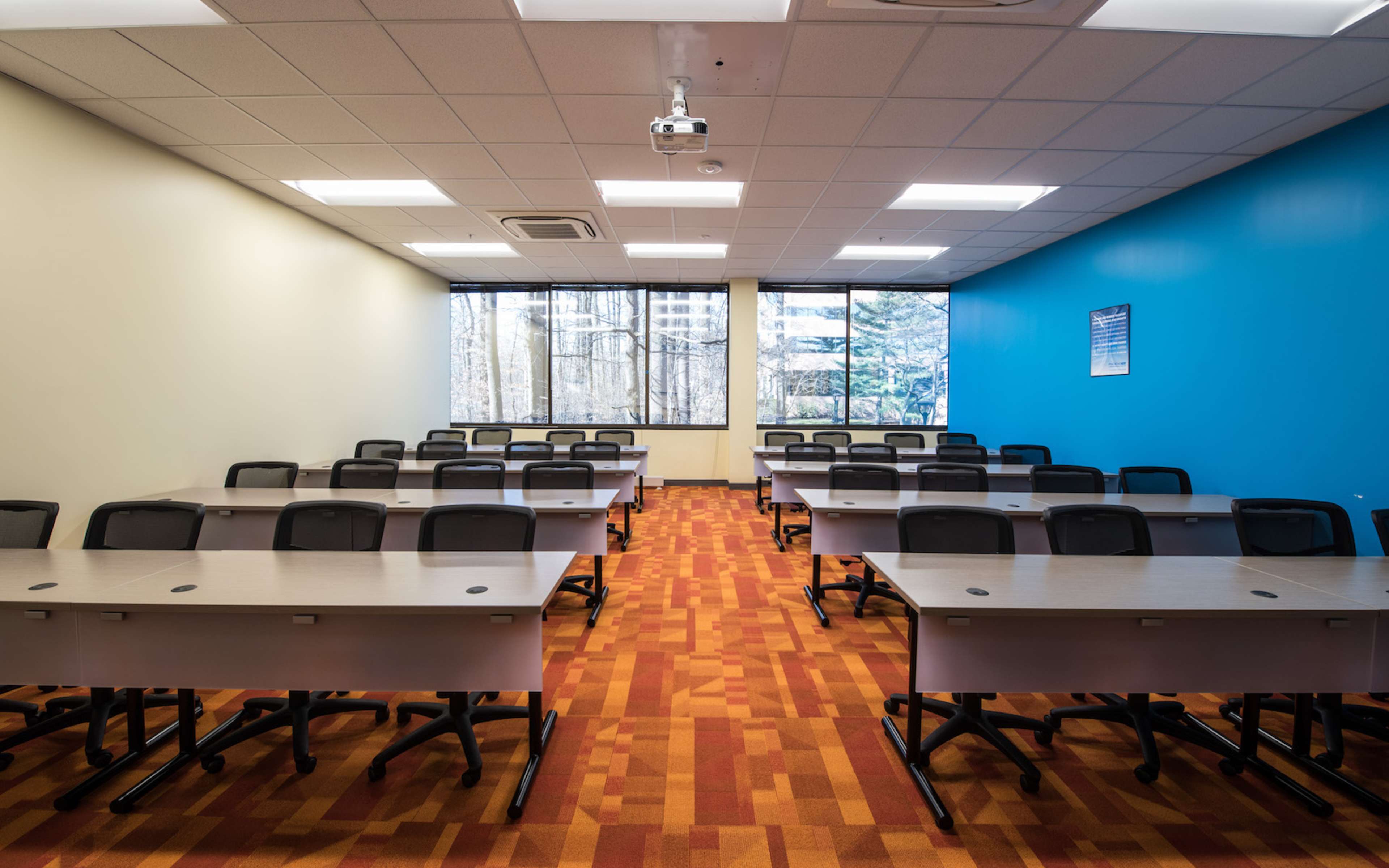 The image shows a modern classroom with several rows of desks and chairs arranged neatly, featuring large windows that allow natural light to enter.
