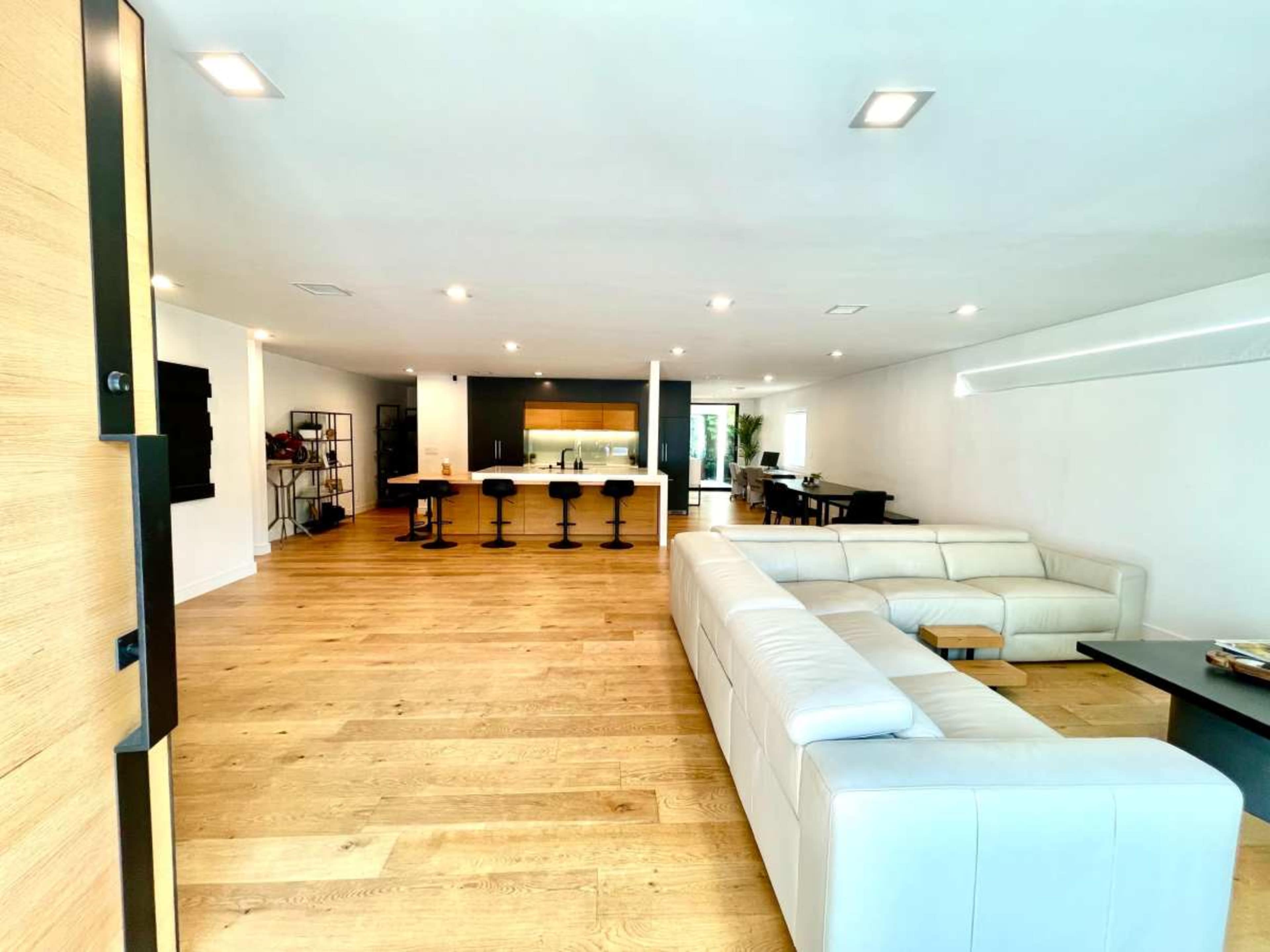 The image shows a spacious, modern living area featuring a white leather sectional sofa, a dining table, and a kitchen with bar stools in the background.