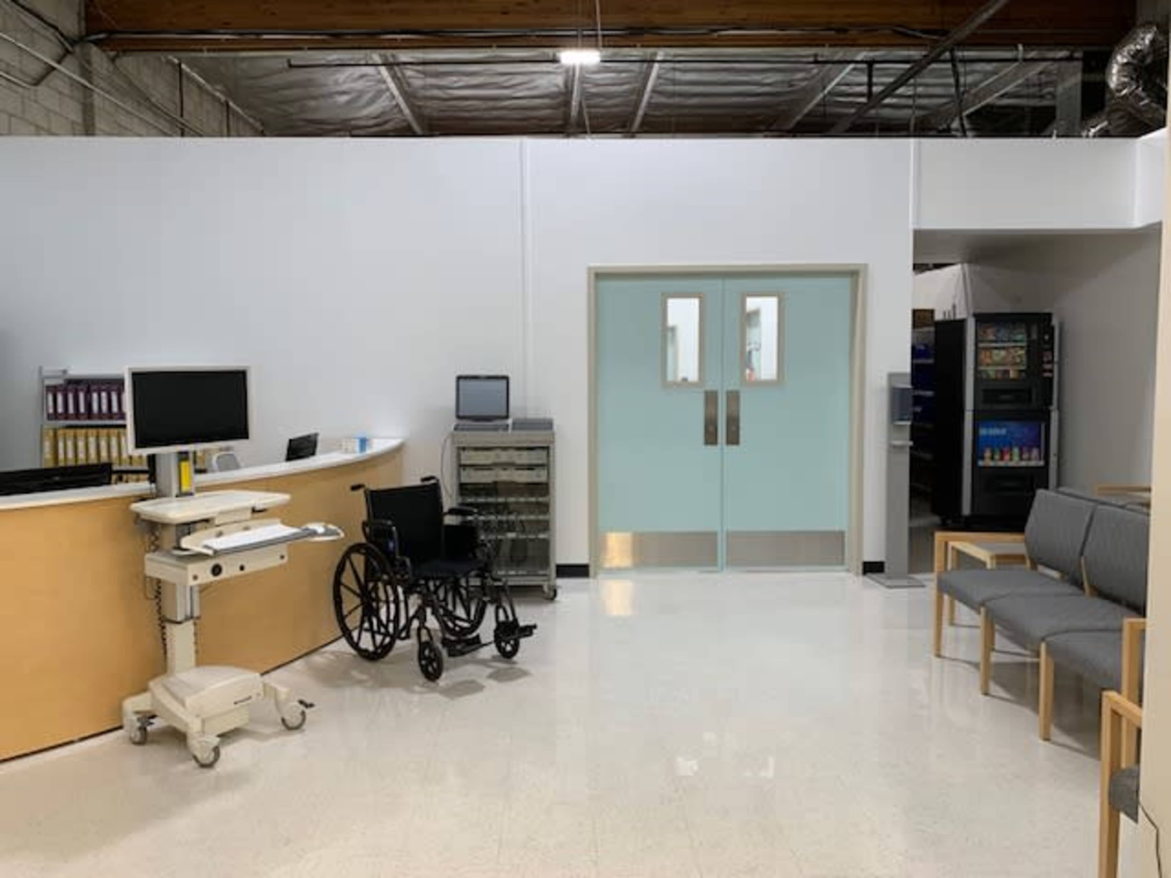 A medical waiting area with a reception desk, a wheelchair, and double doors leading to another space.