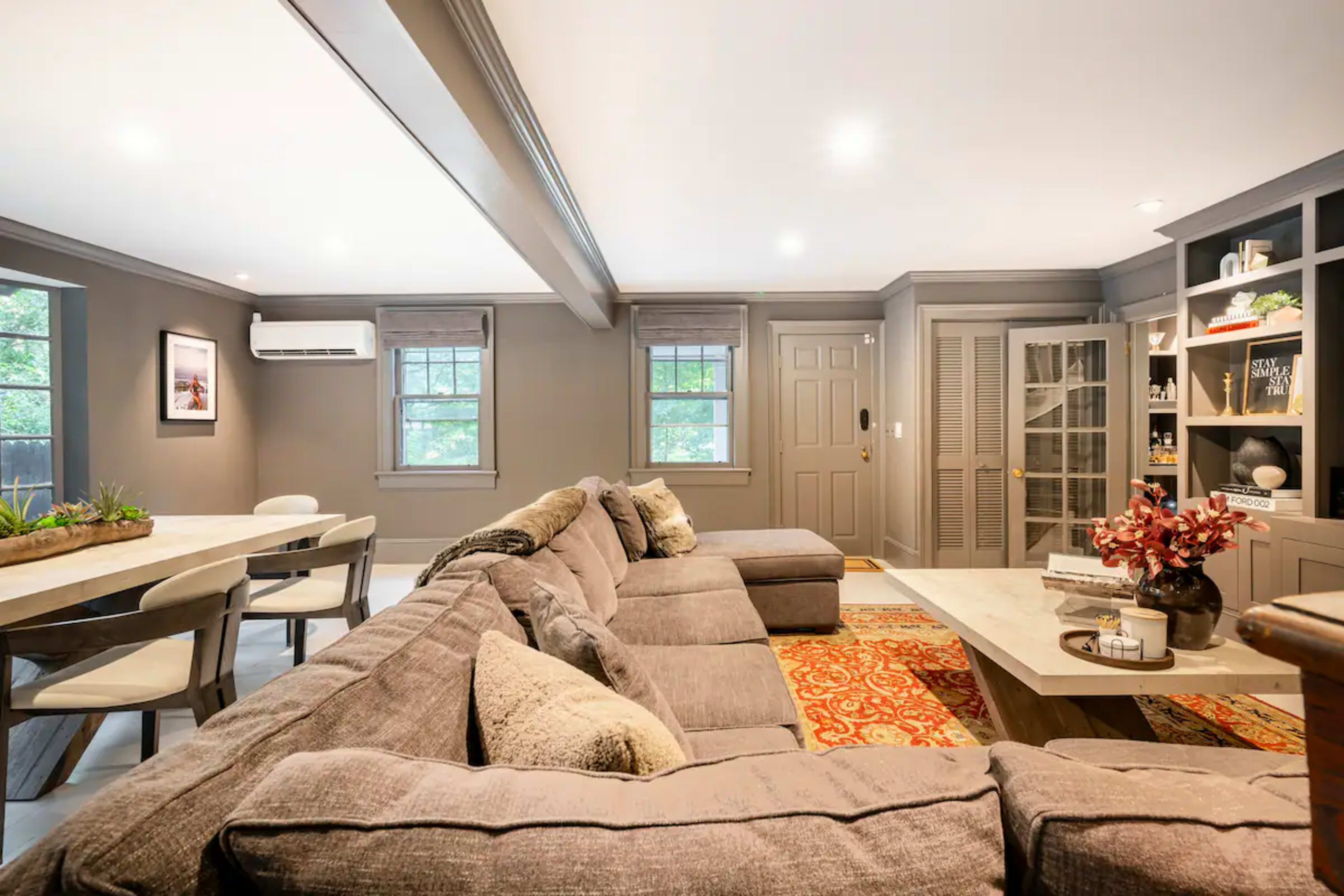 A modern living room features a large sectional sofa, a wooden dining table, and built-in shelves with decorative items.
