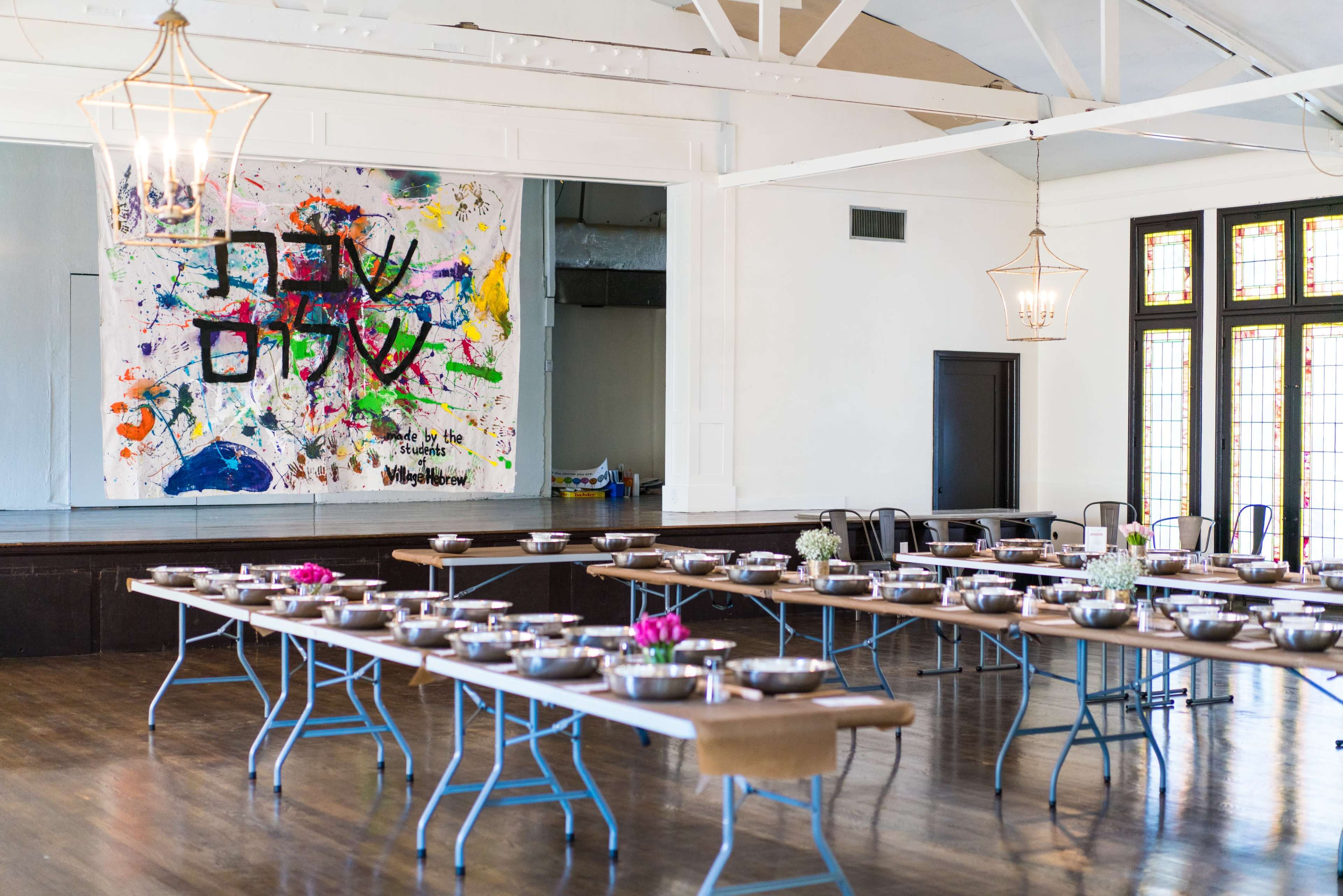 A large room features tables arranged in rows, each set with bowls and small flower arrangements, alongside a colorful abstract mural on the wall.