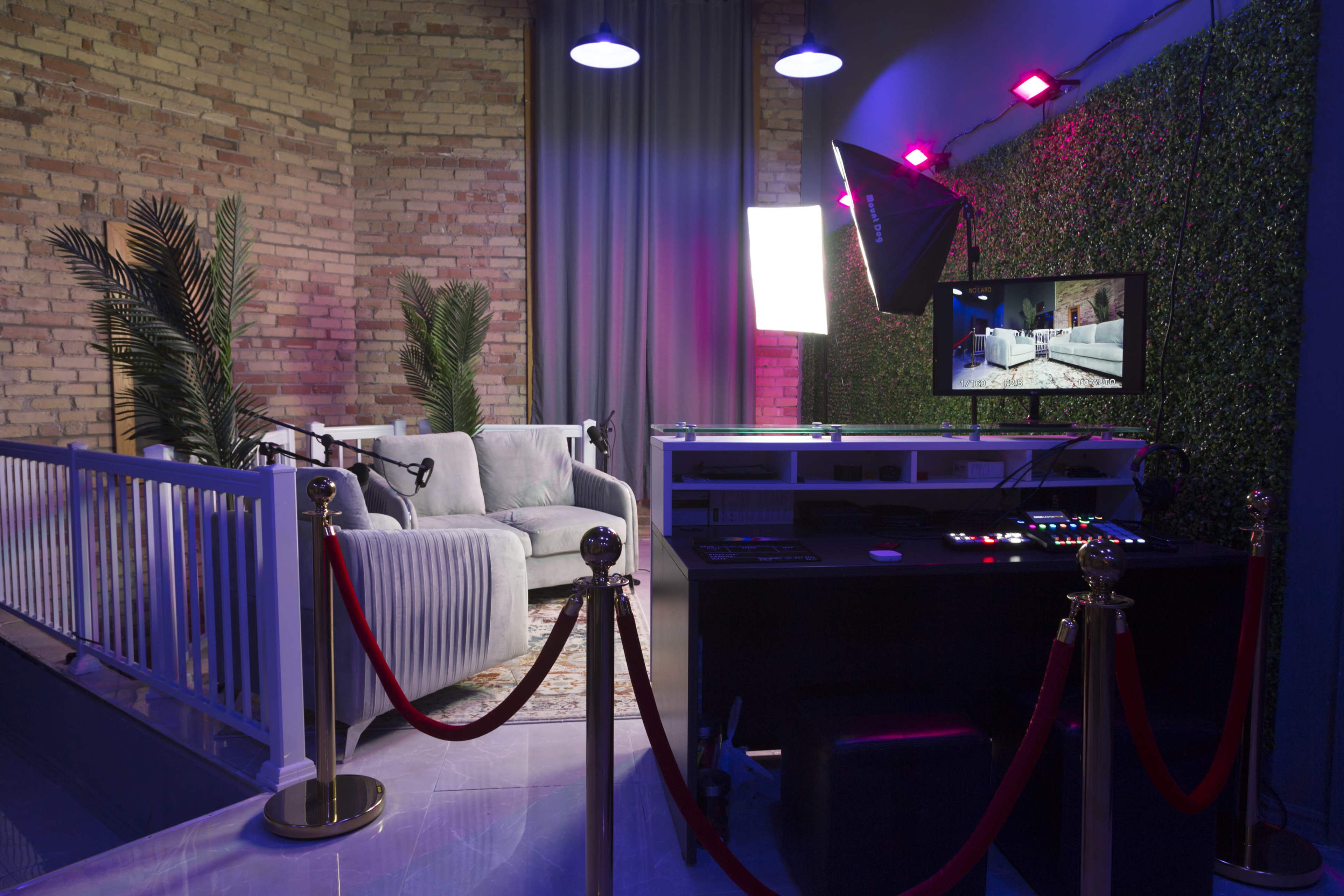 A media production studio featuring a cozy seating area with a backdrop of greenery and colorful lighting, along with a control desk and monitor.