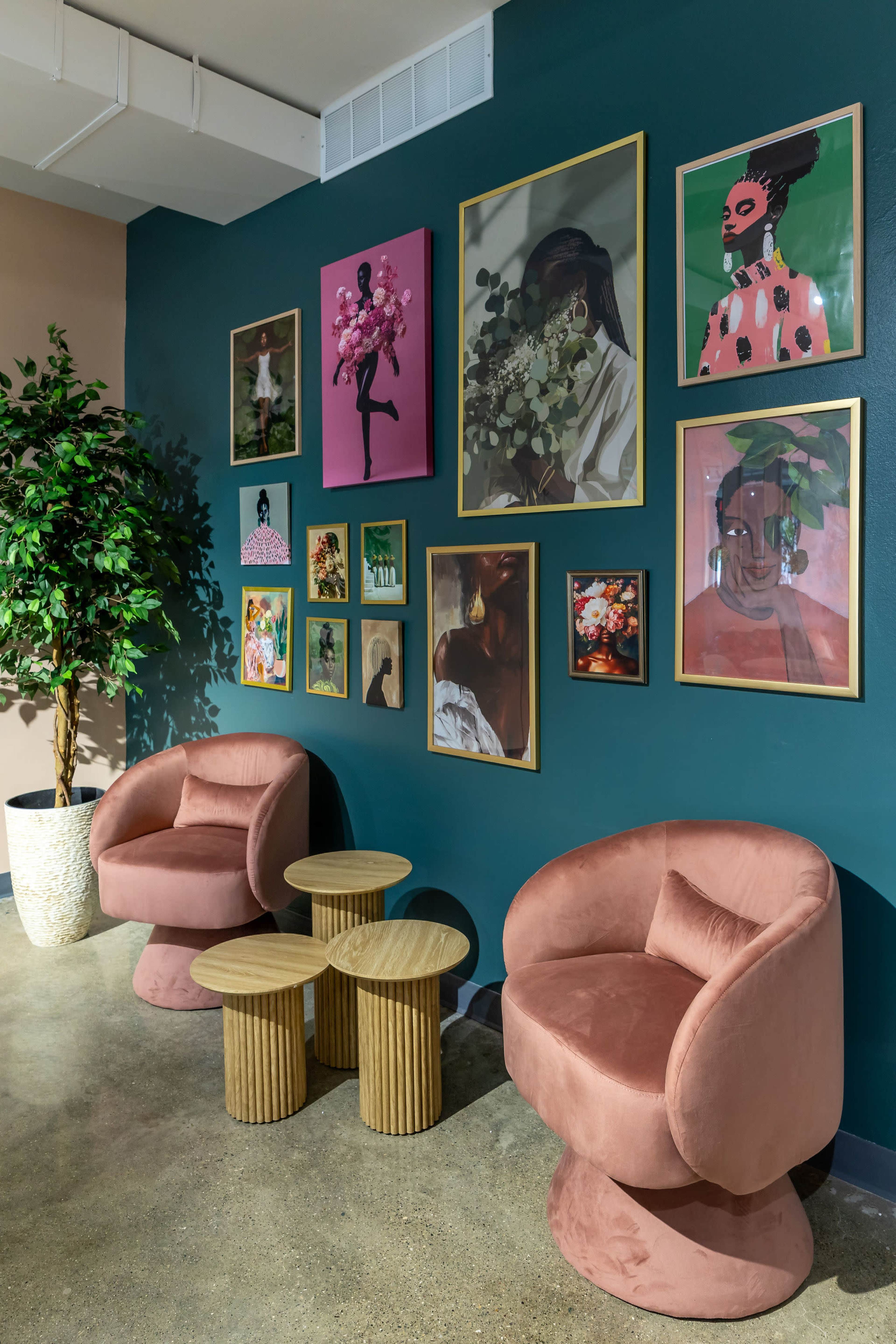 The image shows a seating area with two pink, round chairs, a set of three cylindrical wooden tables, and a wall decorated with various framed artworks on a teal background.