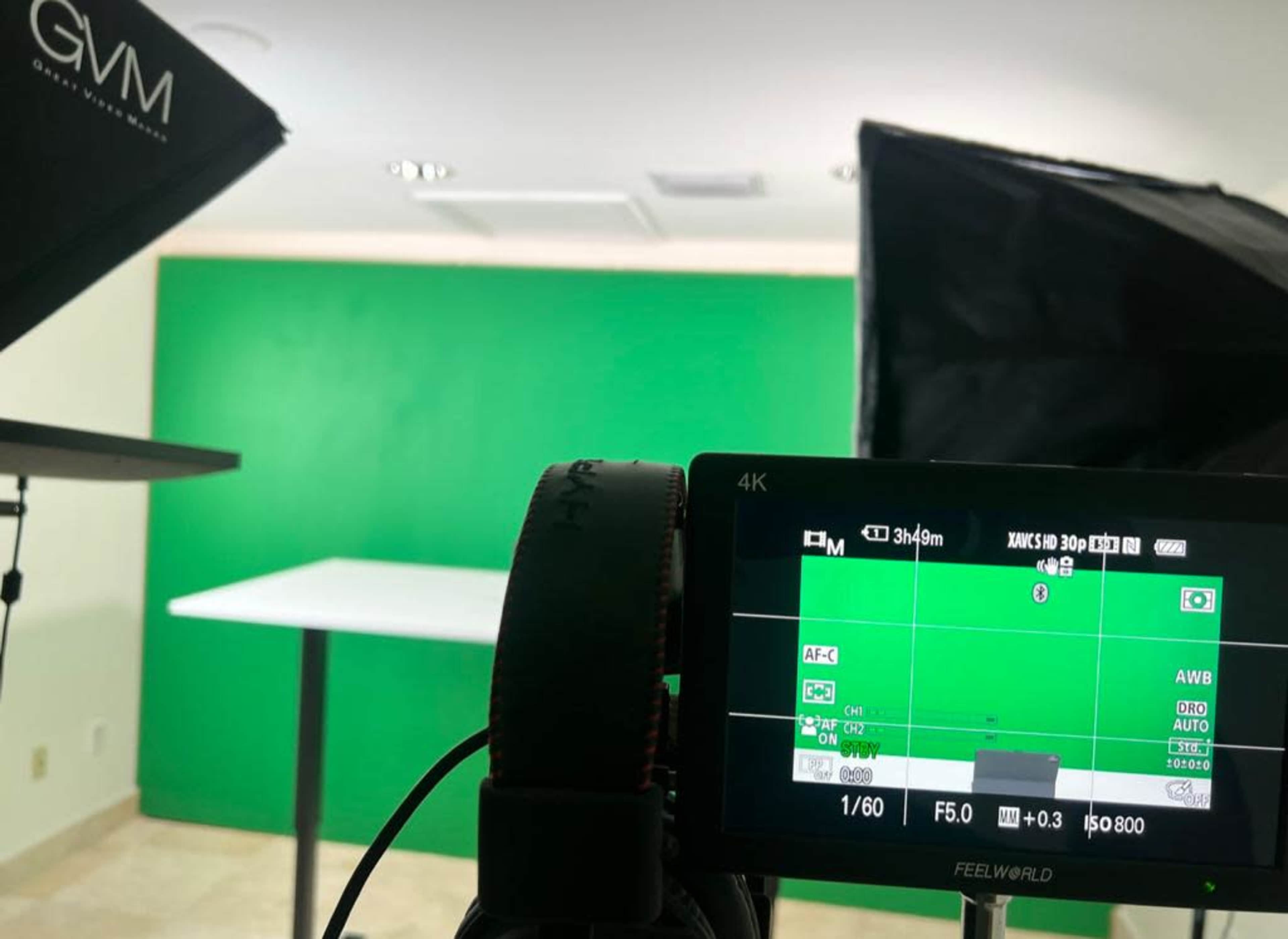 A camera setup with a monitor displaying a live view of a green screen and a table, with lighting equipment positioned on either side.