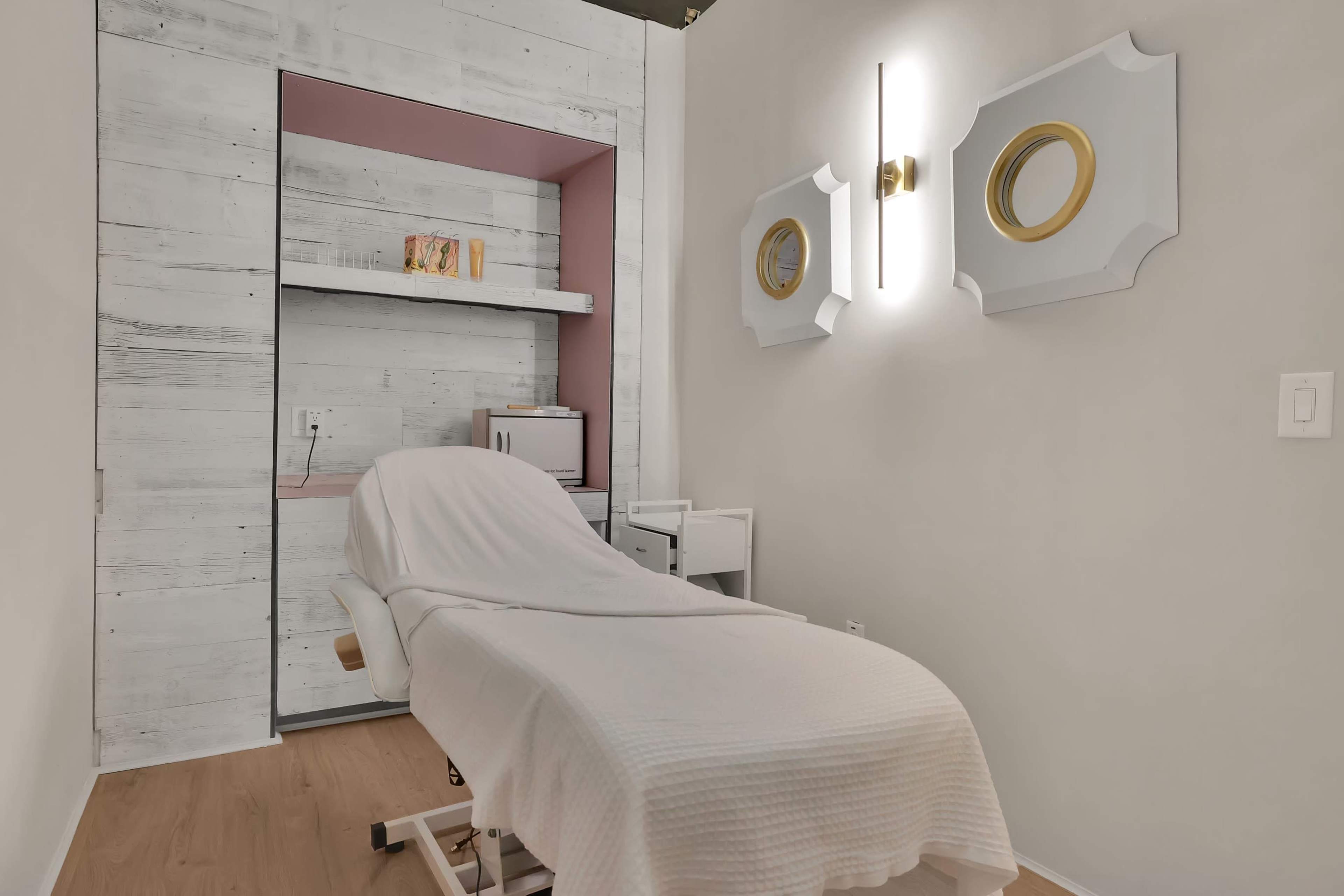 The image shows a treatment room featuring a therapy bed covered with a white sheet, flanked by modern lighting and decorative mirrors on the walls.