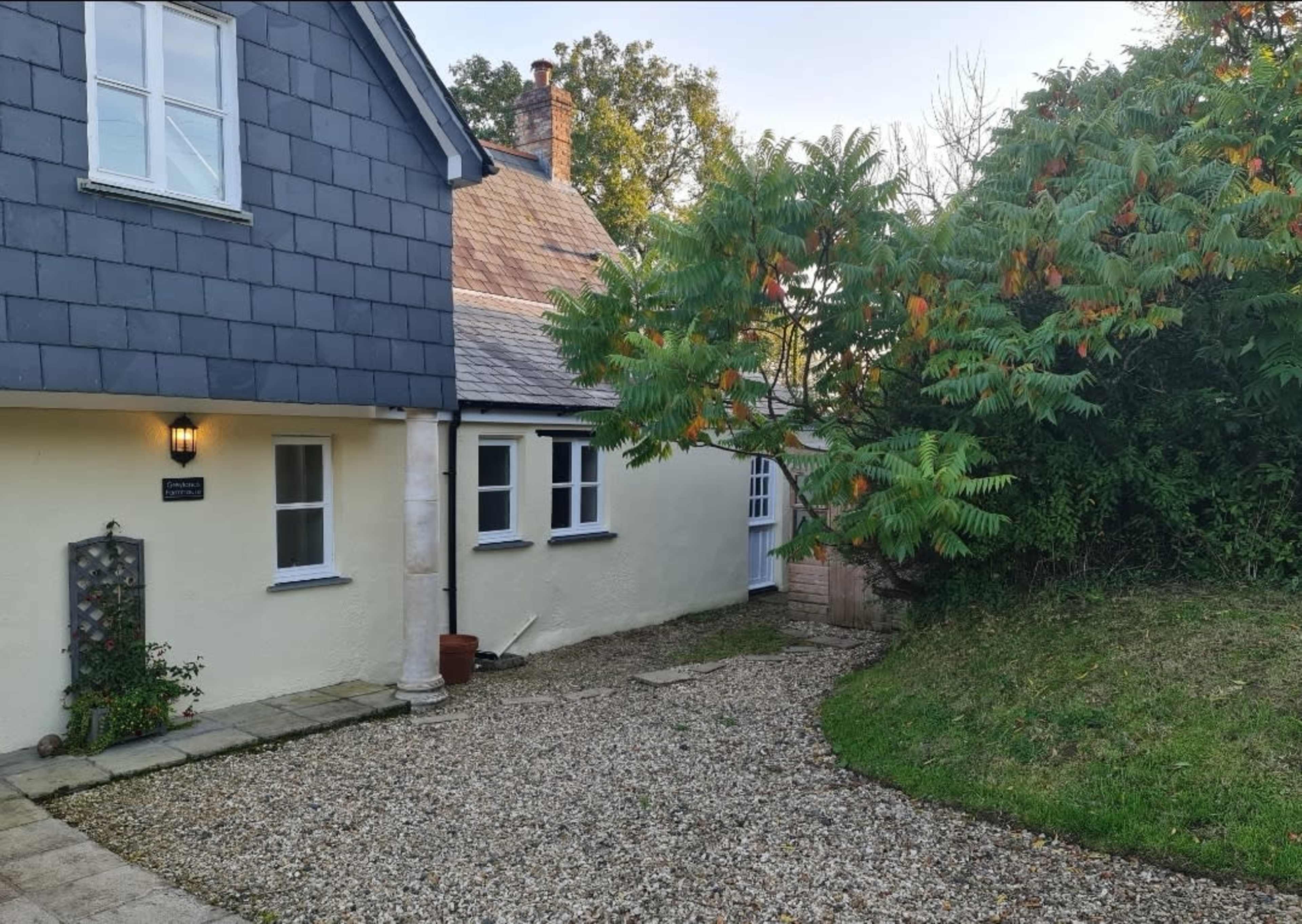 A house with a gravel pathway leading to a door, surrounded by green foliage and trees.