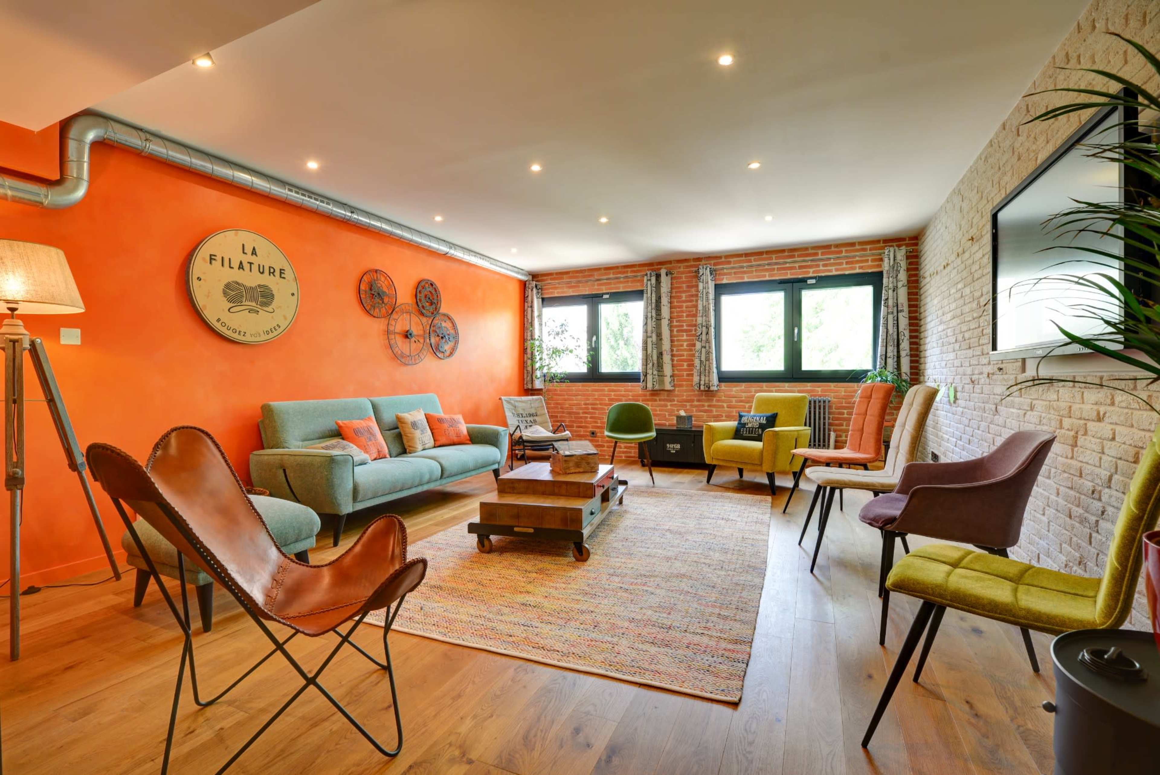 The room features a mix of seating options arranged on a colorful rug, with an orange accent wall and large windows providing natural light.