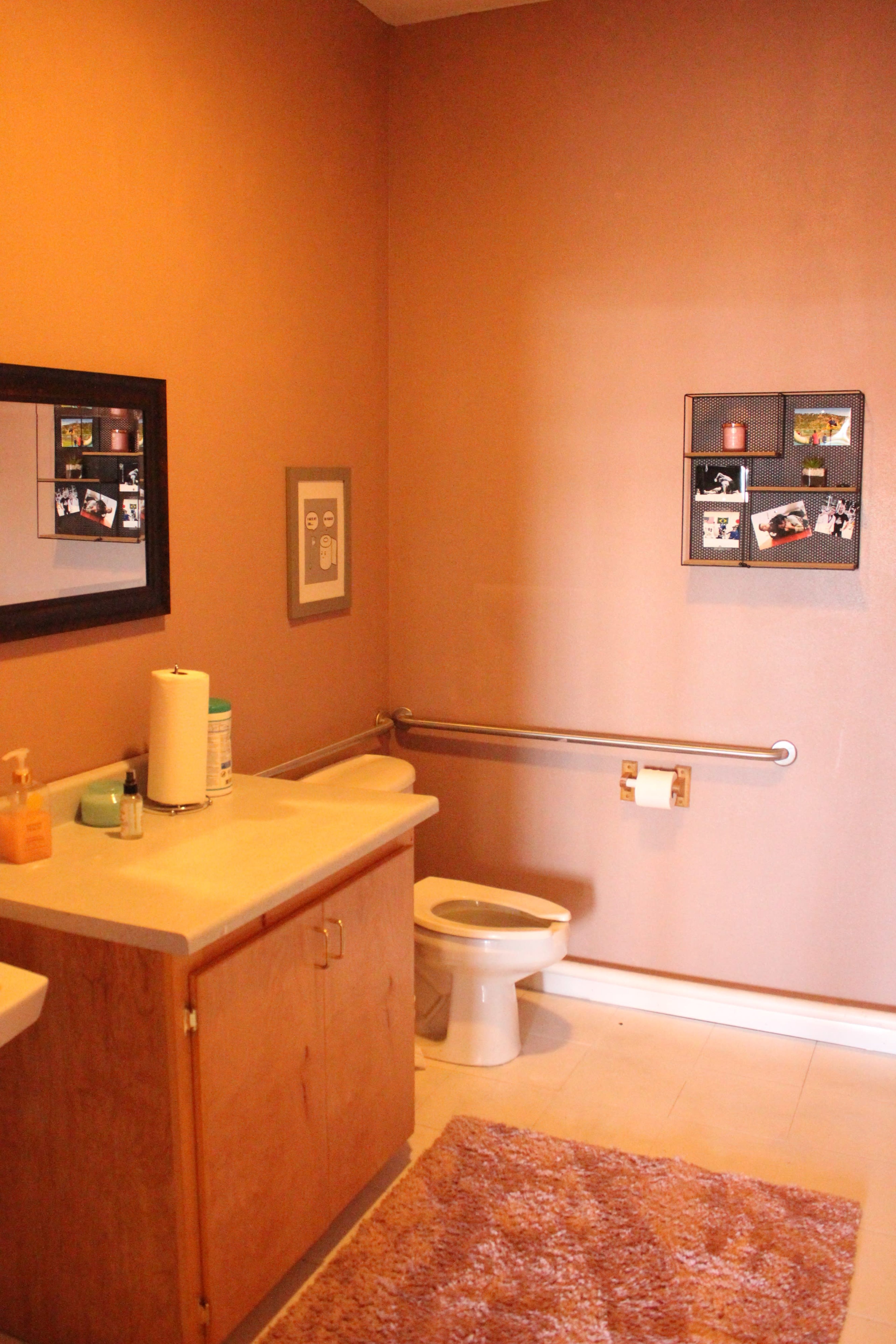 The image shows a bathroom featuring a beige wall, a toilet, a sink with a cabinet, and framed photos mounted on the wall.