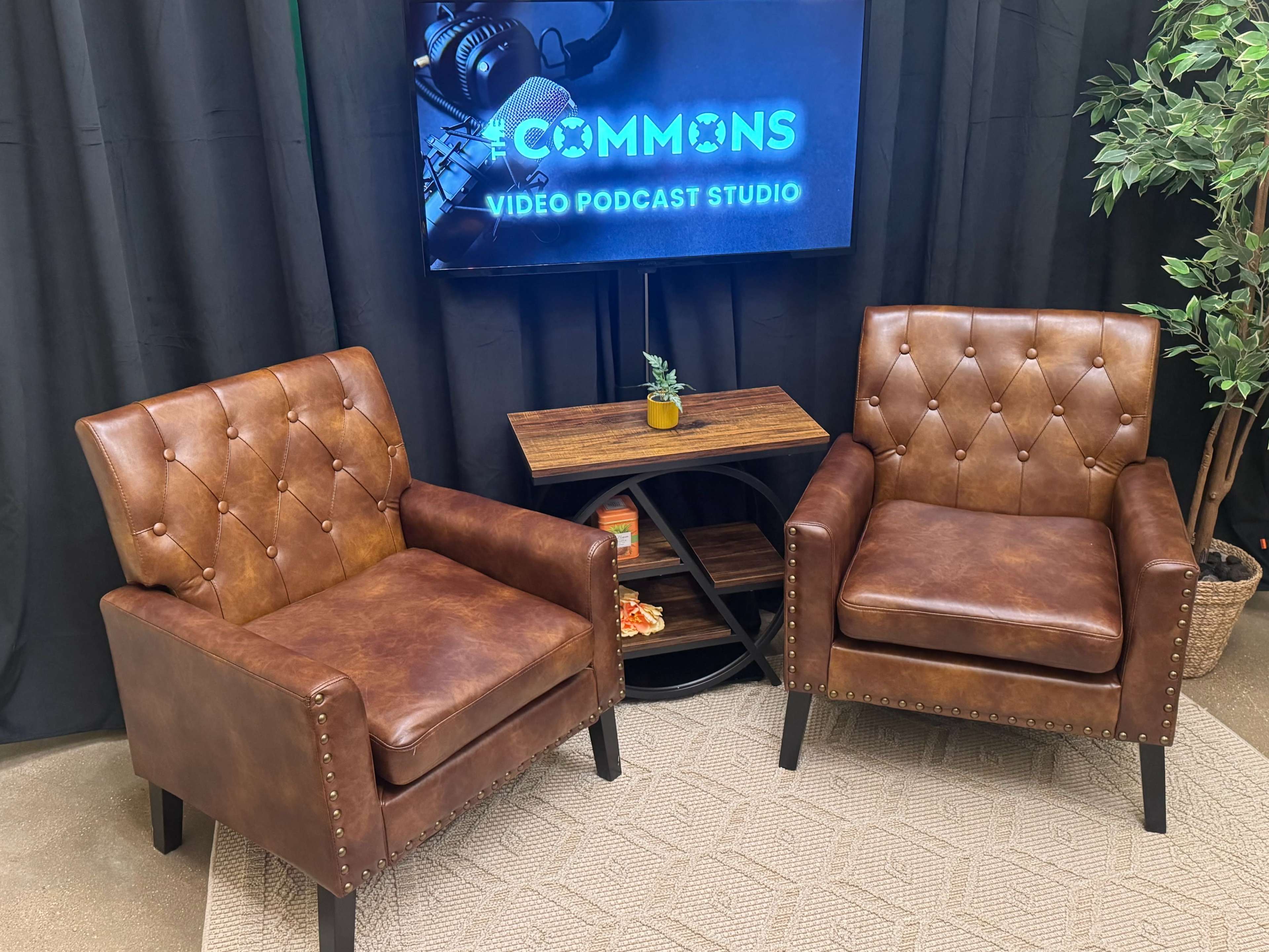 The image shows a video podcast studio setup featuring two brown upholstered chairs facing a small wooden table, with a screen displaying "COMMONS VIDEO PODCAST STUDIO" in the background.