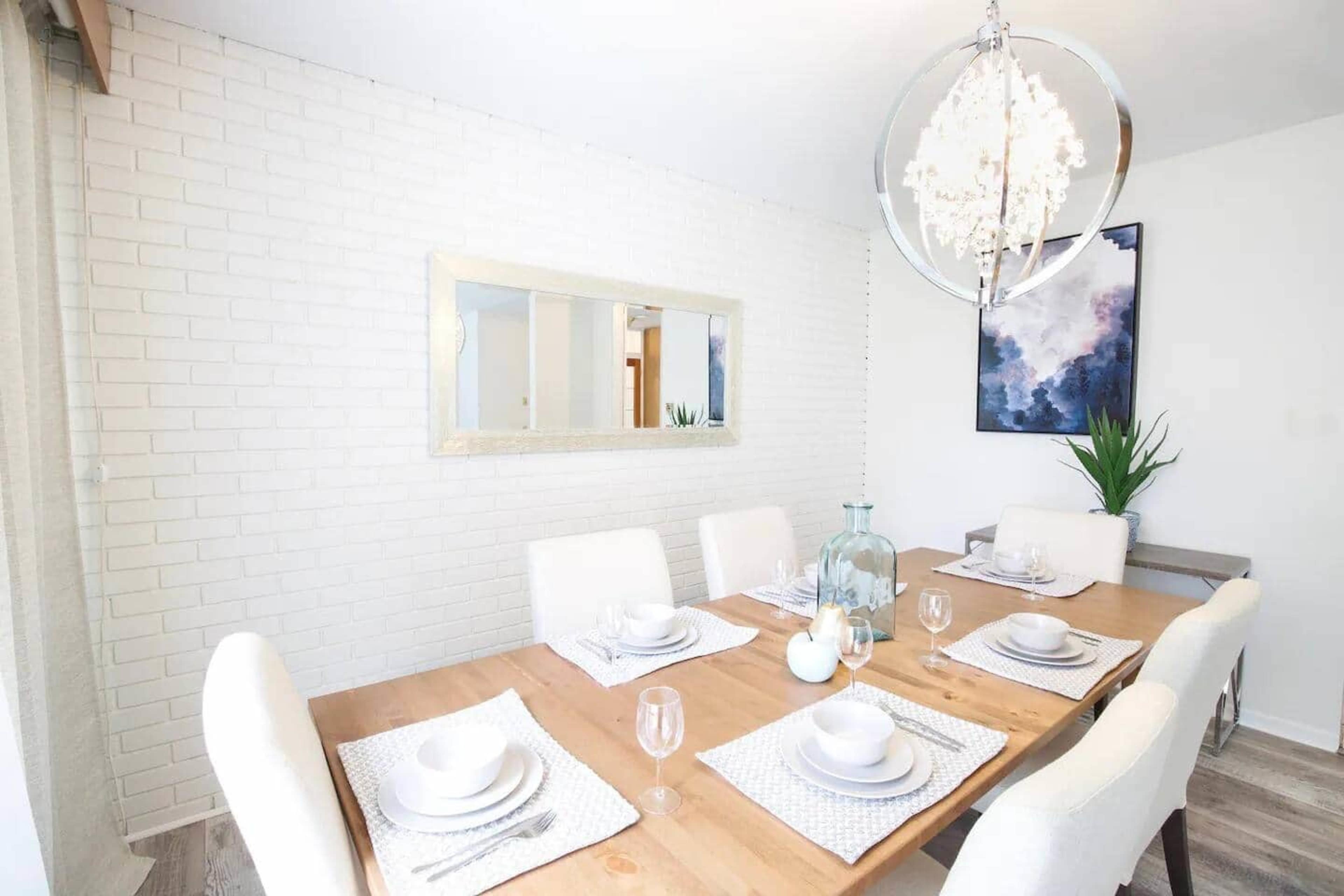 The image shows a modern dining room with a wooden table set for dinner, featuring white plates, glasses, and a decorative centerpiece, against a backdrop of a white brick wall and contemporary art.