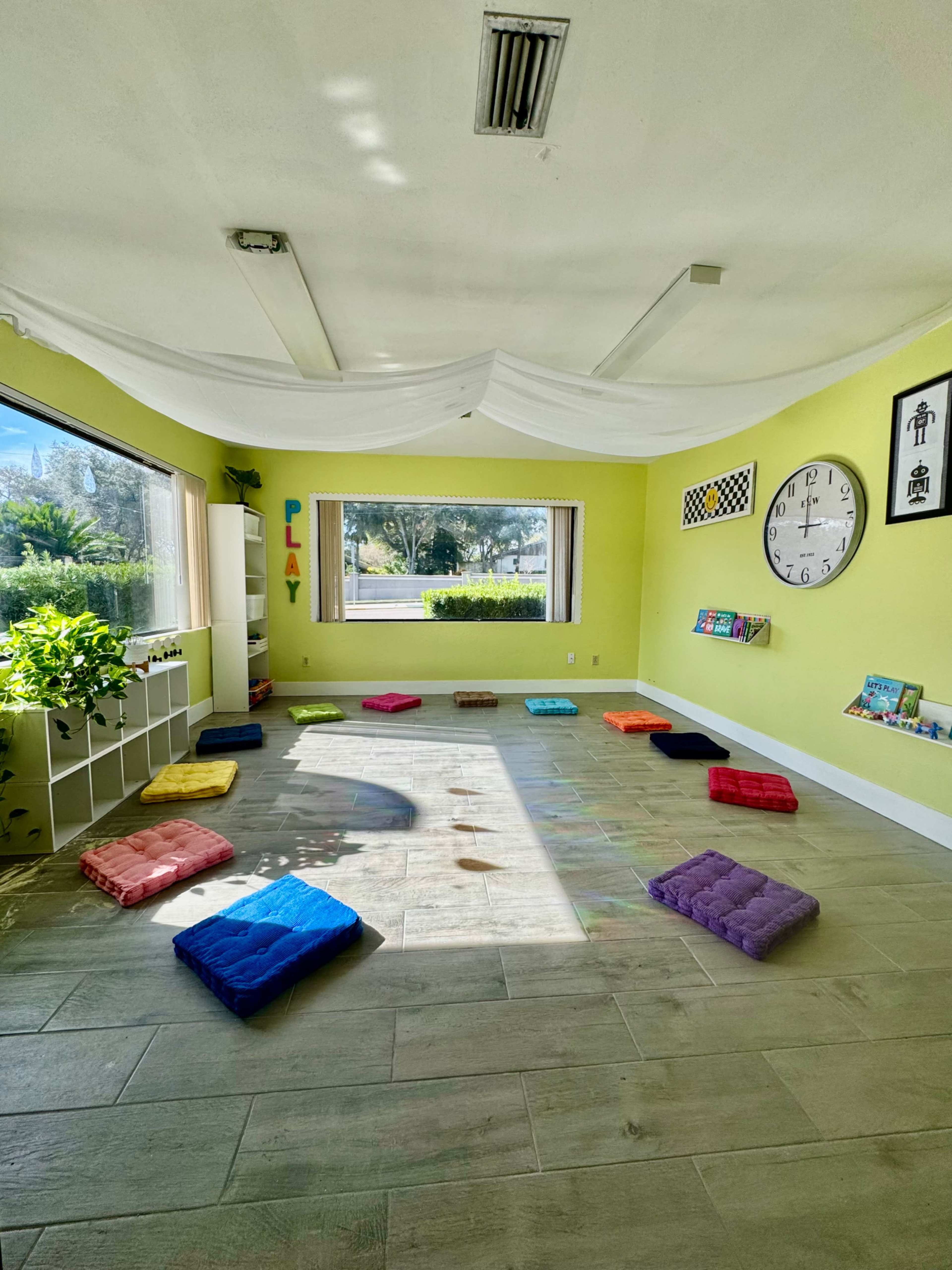 The image shows a brightly lit room with green walls, several colorful cushions scattered on the floor, and large windows offering a view of the outdoors.