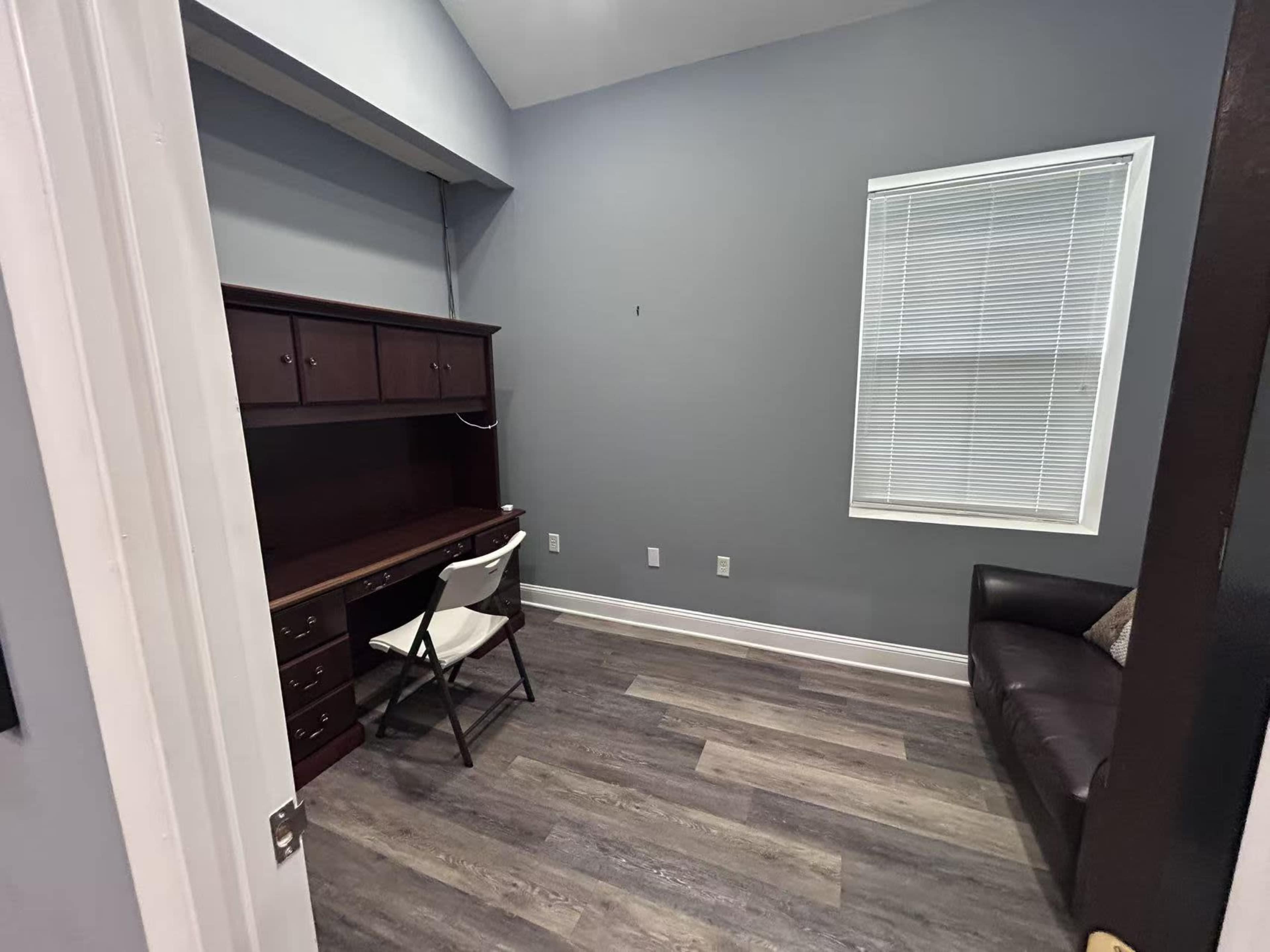 The image shows a small, semi-furnished room with a dark wooden desk, a white chair, and a brown sofa, along with a window that allows natural light.