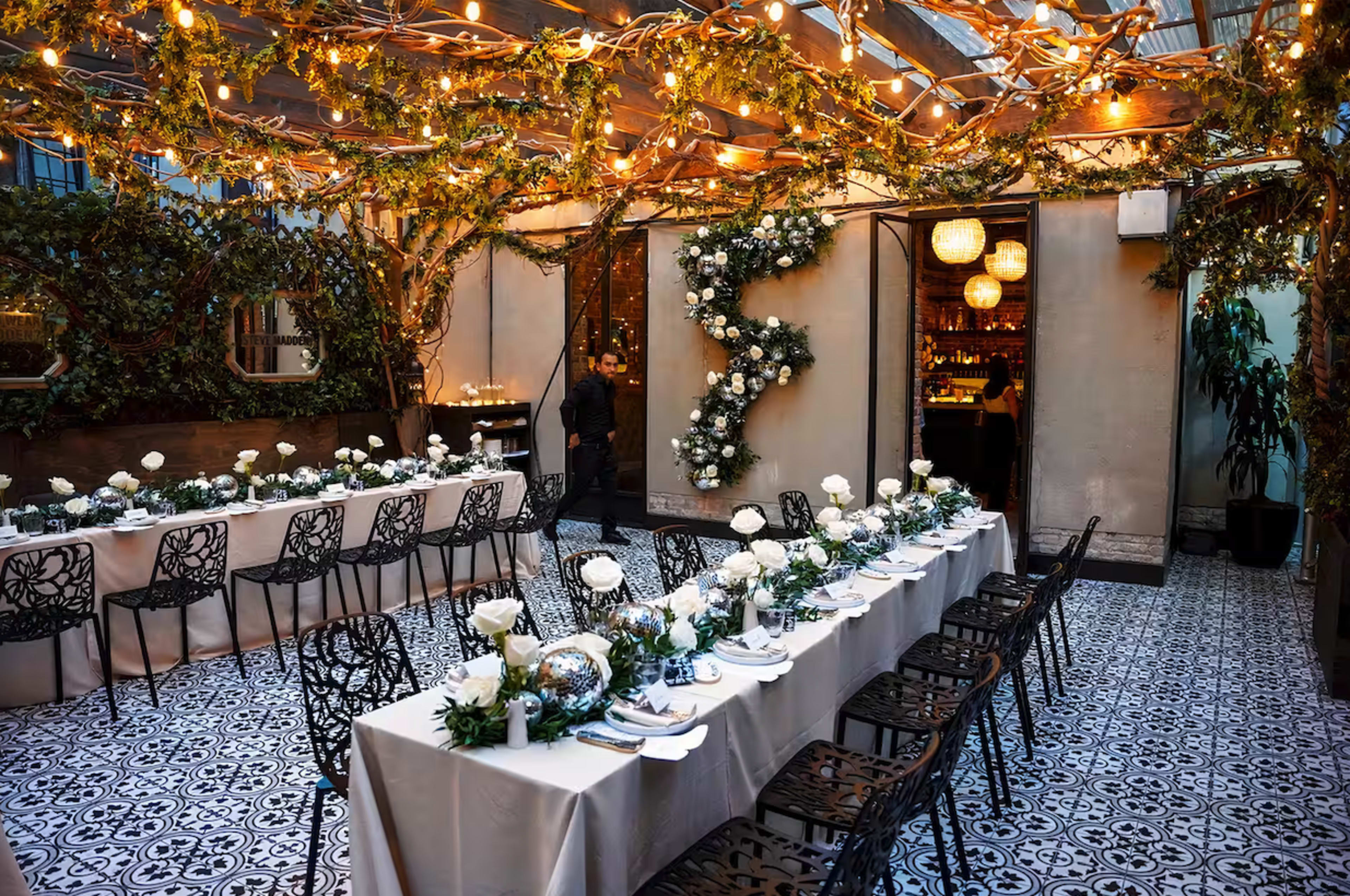The image shows a beautifully decorated indoor dining area with long tables set for a meal, surrounded by greenery and string lights.