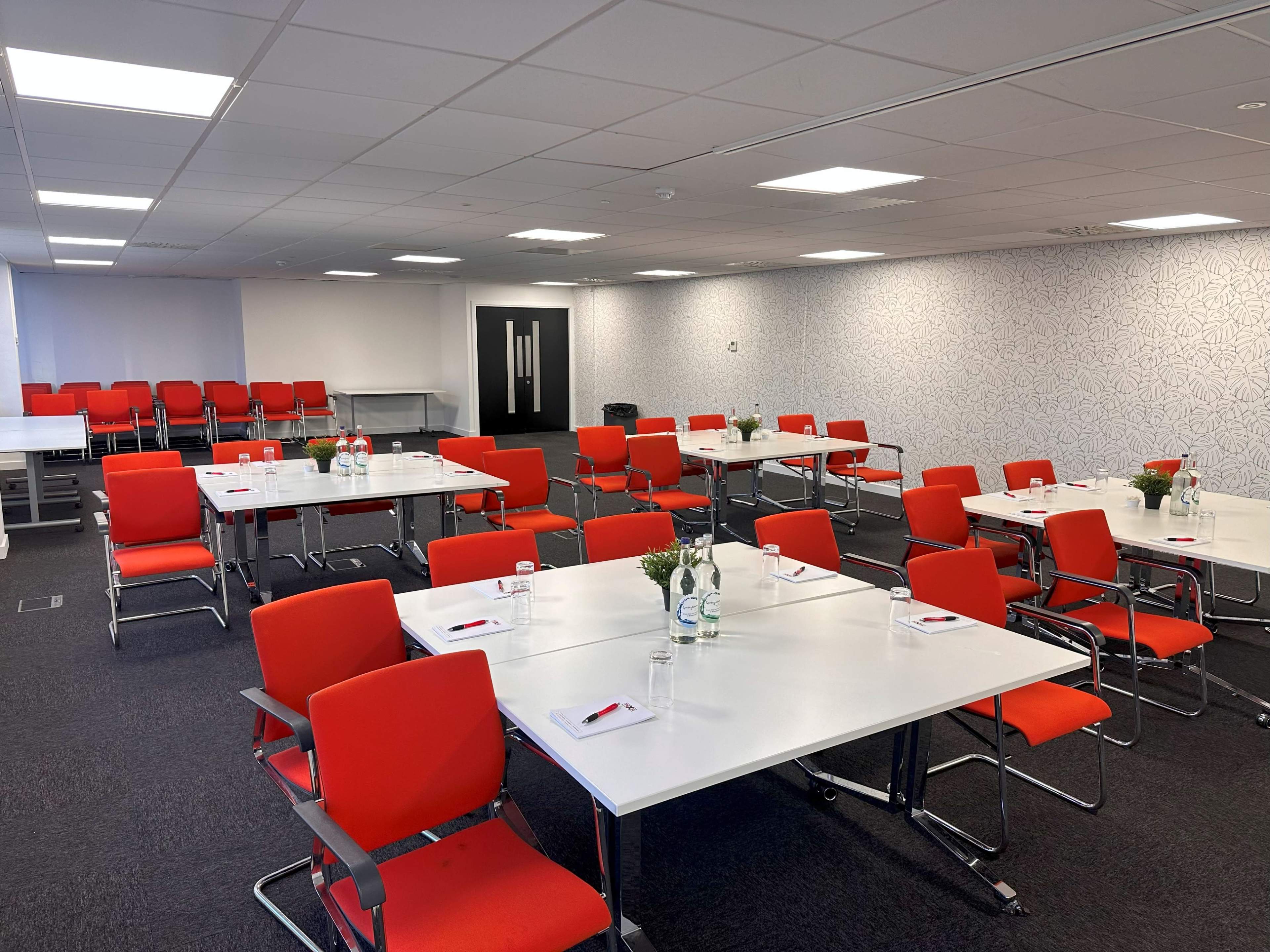 A modern meeting room features several white tables arranged with red chairs and small floral centerpieces.