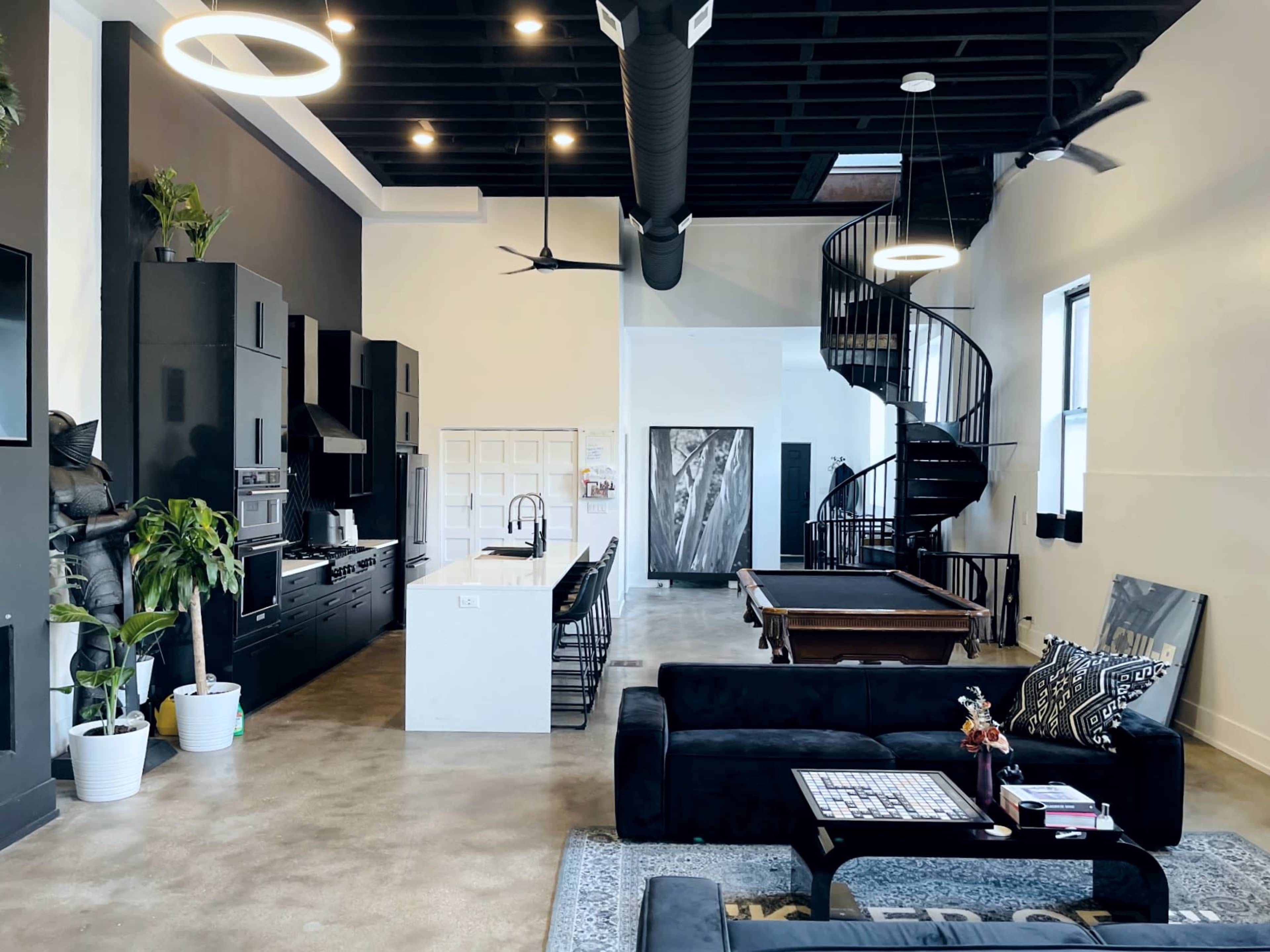 A modern open-concept living space features a kitchen with black cabinetry, a spiral staircase, and a pool table adjacent to a seating area with a black sofa and decorative elements.