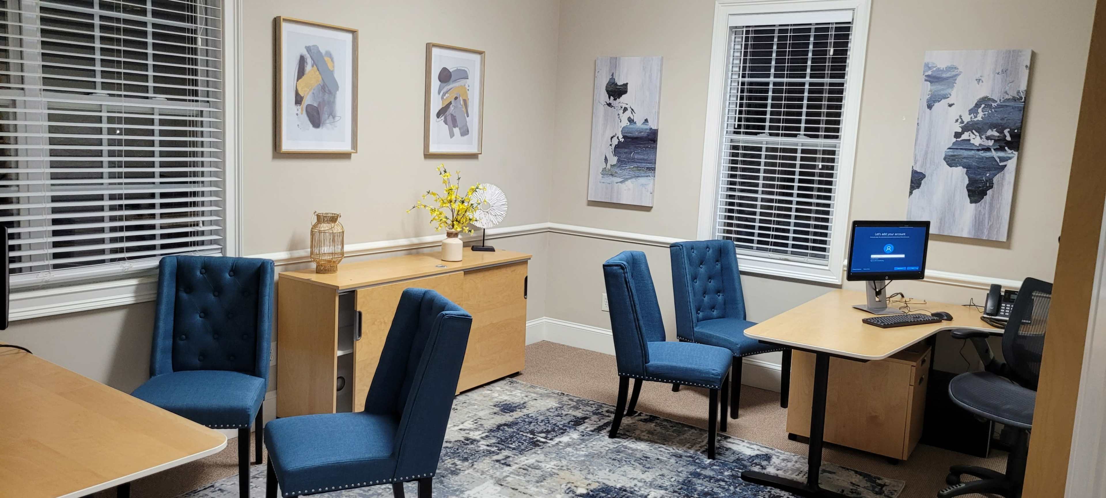 The image shows a small office space with blue chairs, a wooden desk and cabinets, and framed artwork on the walls.