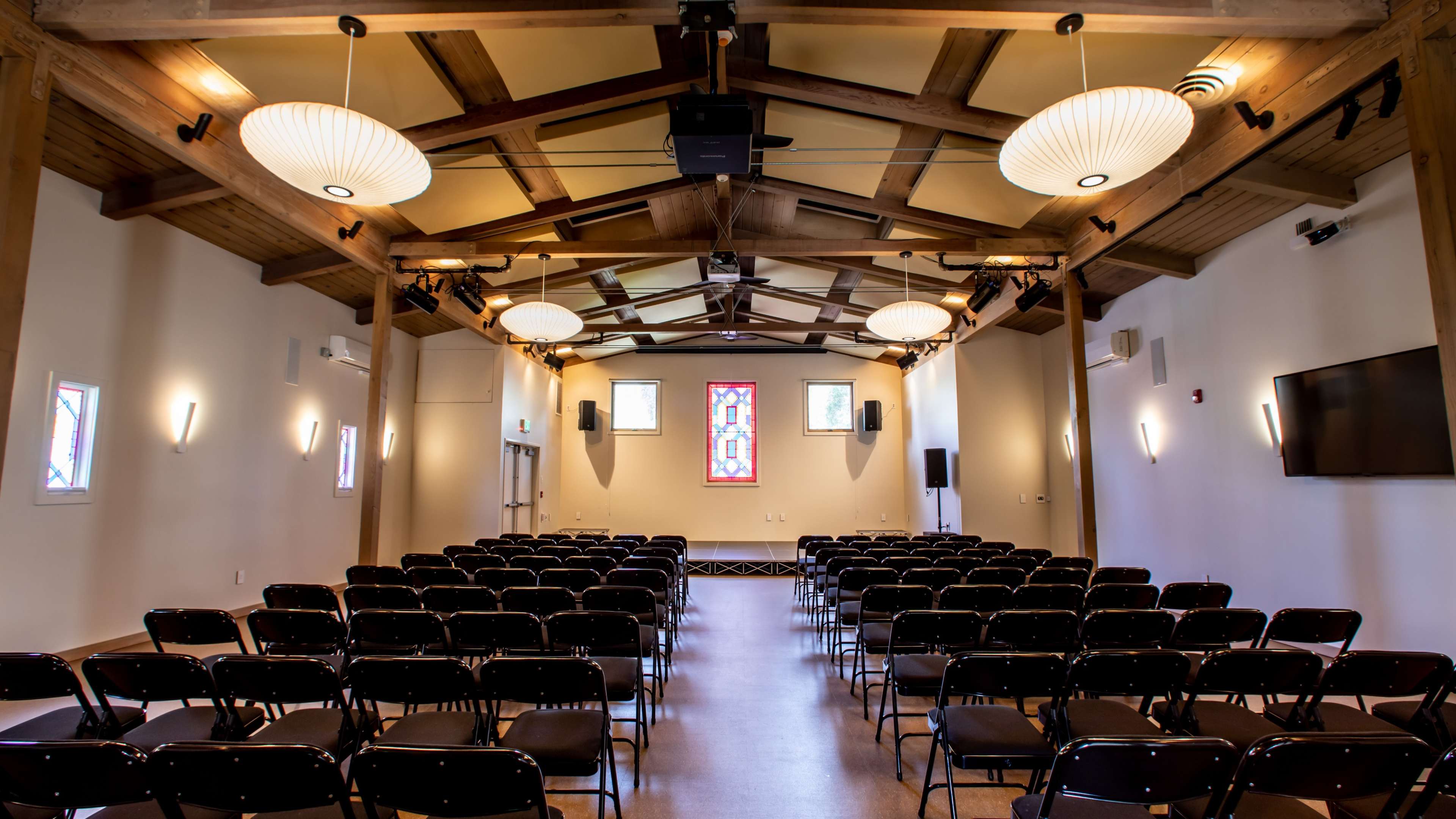 A spacious indoor hall with rows of black chairs facing a stained glass window and a wall-mounted screen.