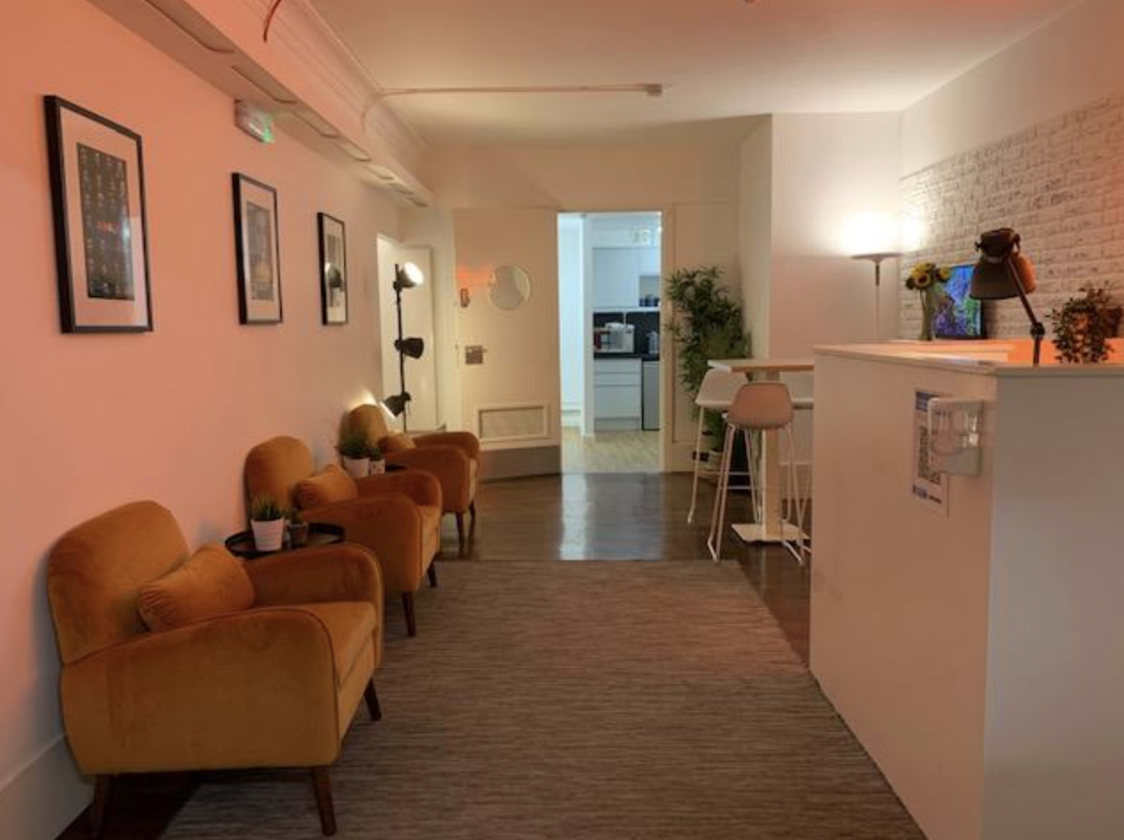 The image shows a well-lit hallway with orange armchairs on one side, a small table, and a doorway leading to a kitchen area in the background.