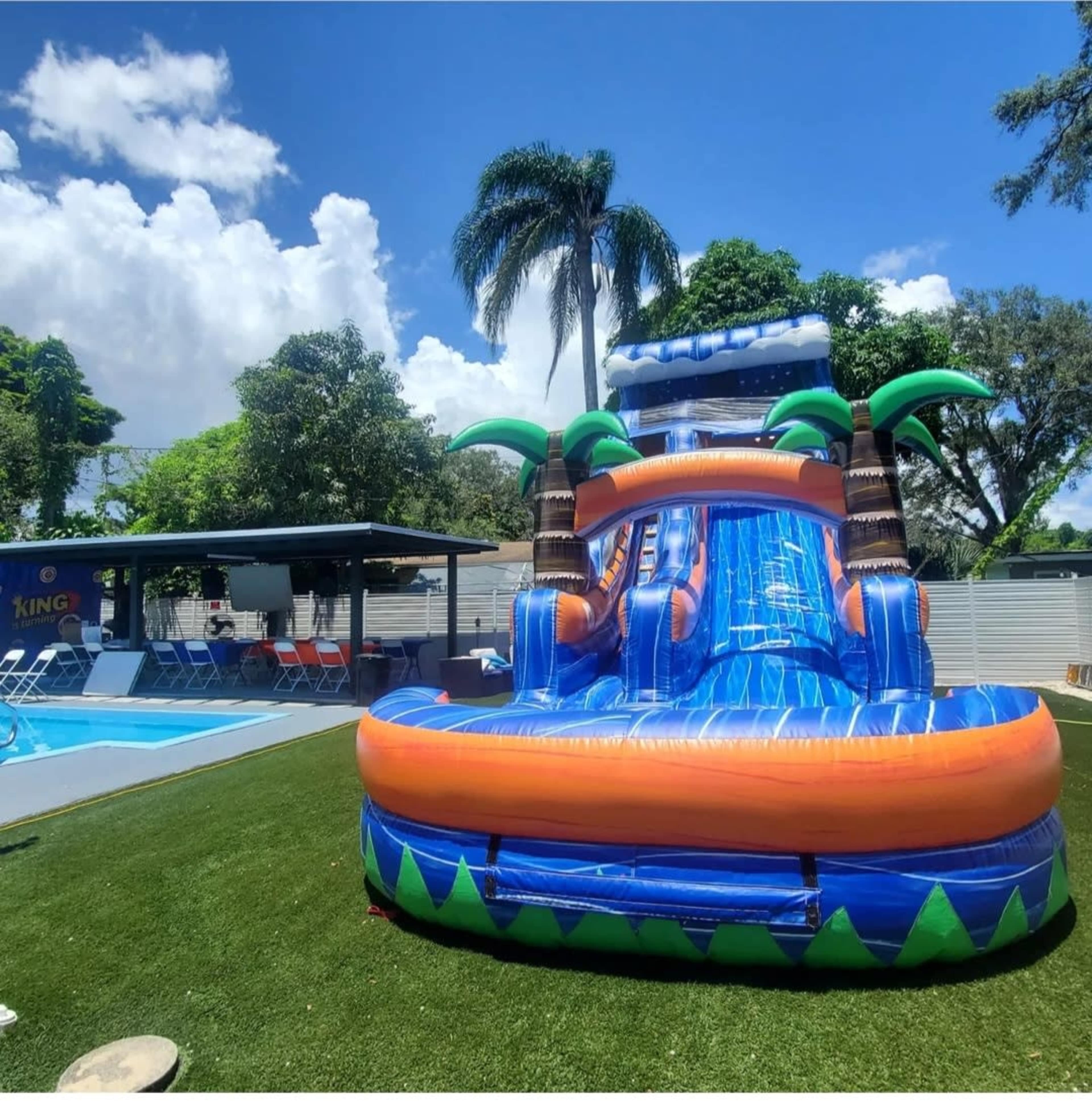 An inflatable water slide shaped like a tropical island is set up near a swimming pool surrounded by lounge chairs and trees.