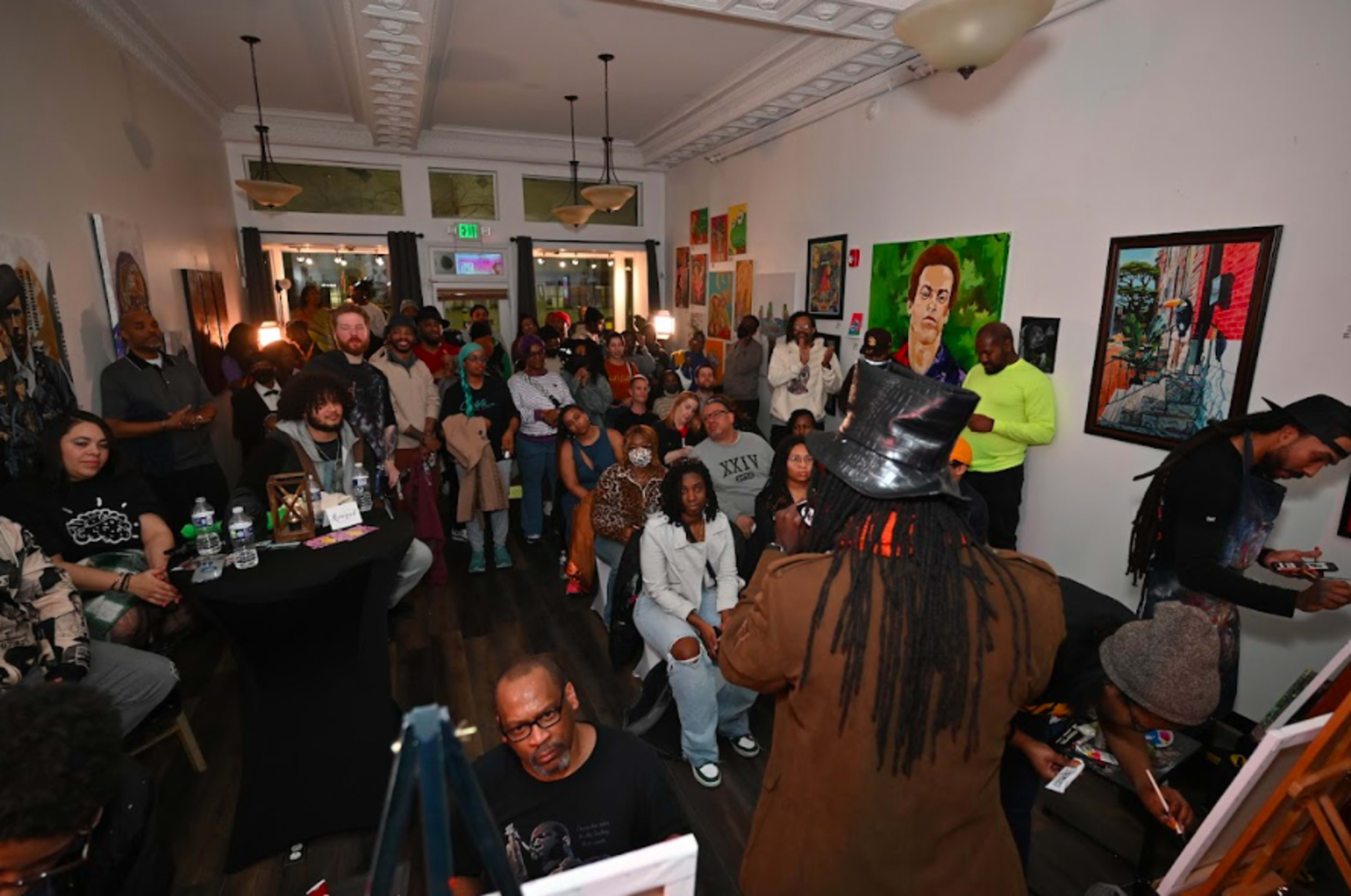 A diverse group of people is gathered in a lively art gallery, some sitting and others standing, as they engage with a speaker in front of them.