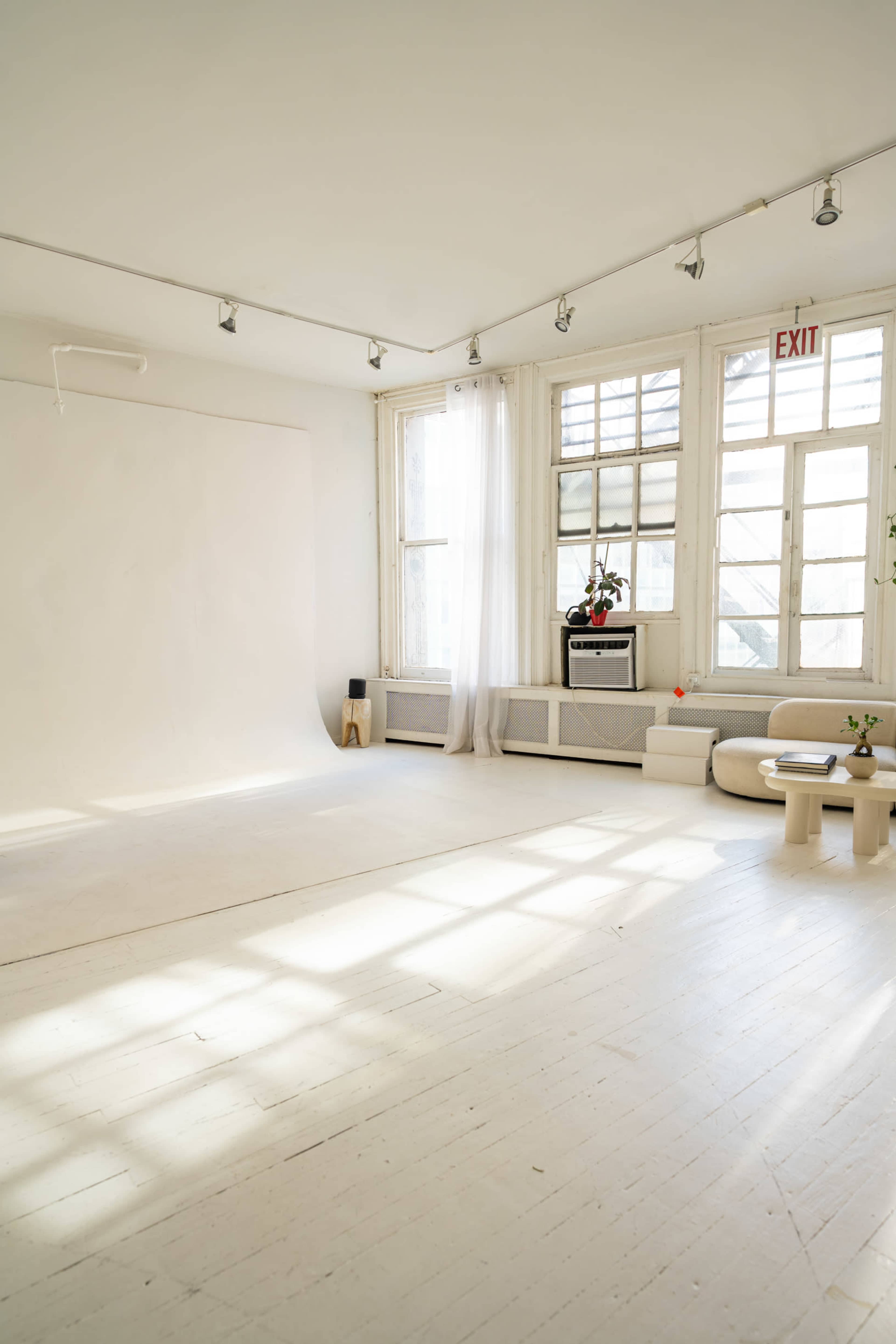The image shows a bright, minimalist studio space with large windows, plain white walls, and wooden flooring.
