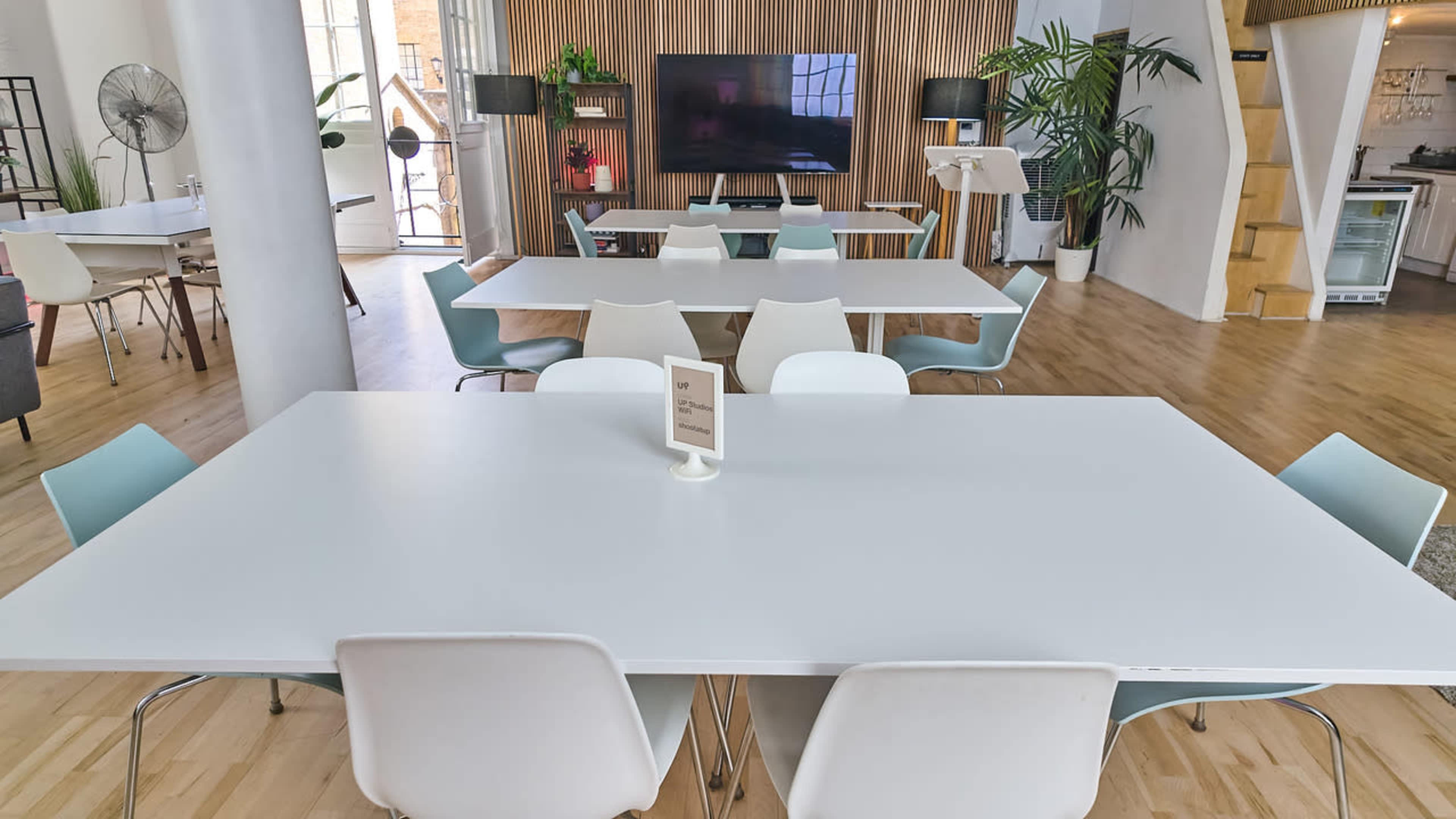 The image shows a modern, minimalist workspace featuring several white tables and chairs arranged in an open area with large windows and decorative plants.