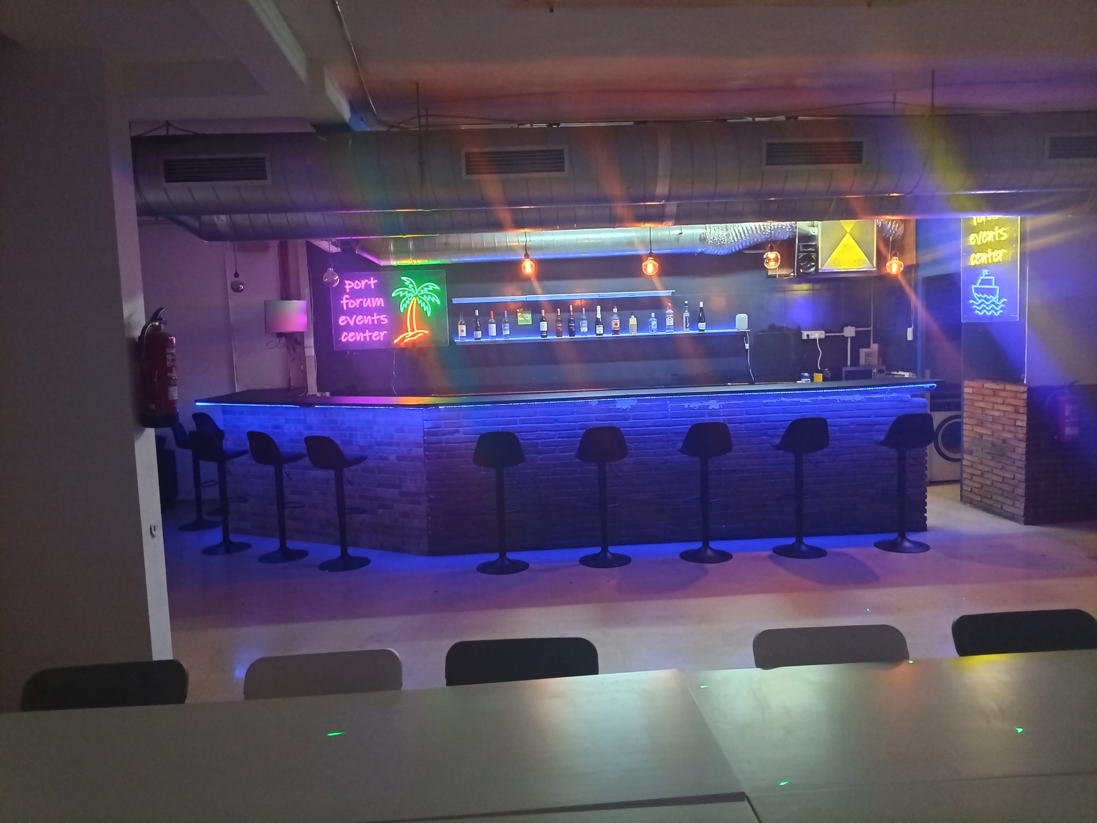The image shows a modern bar area with a blue backlit countertop, high stools, and a neon sign displaying the words "PORT FORUM EVENTS CENTER" alongside decorative elements like a palm tree and a boat.