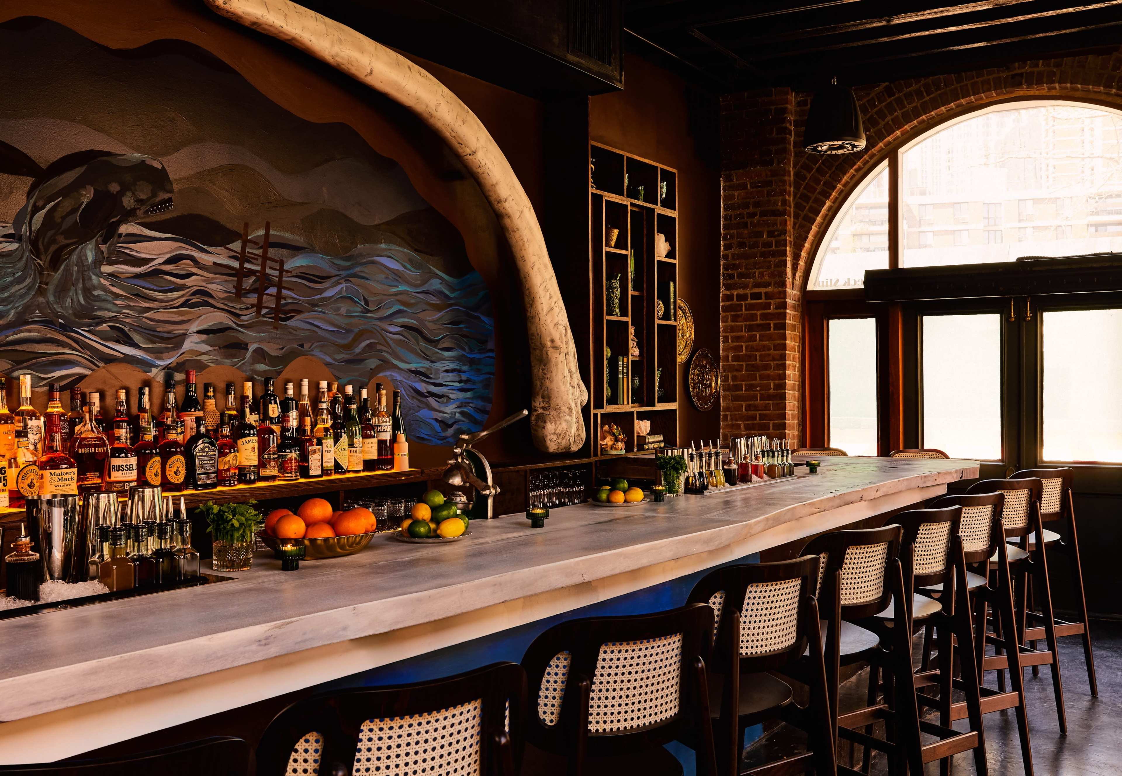 The image shows a bar with a long marble countertop, several bottles of liquor neatly arranged on shelves, and a large mural featuring waves and a whale skull.