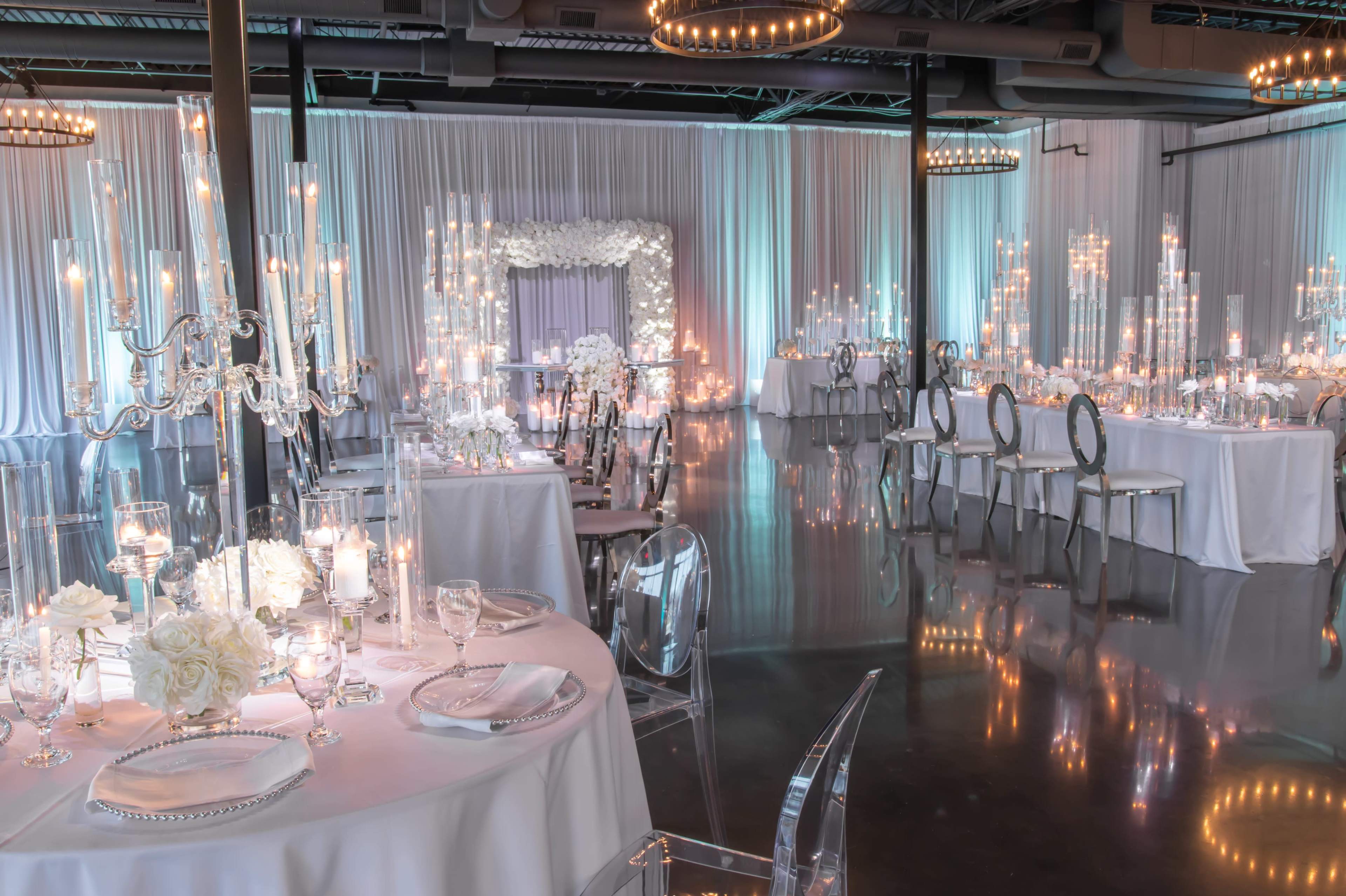 An elegantly decorated event space with tables set for a formal gathering, featuring white tablecloths, floral centerpieces, and ambient candlelight.