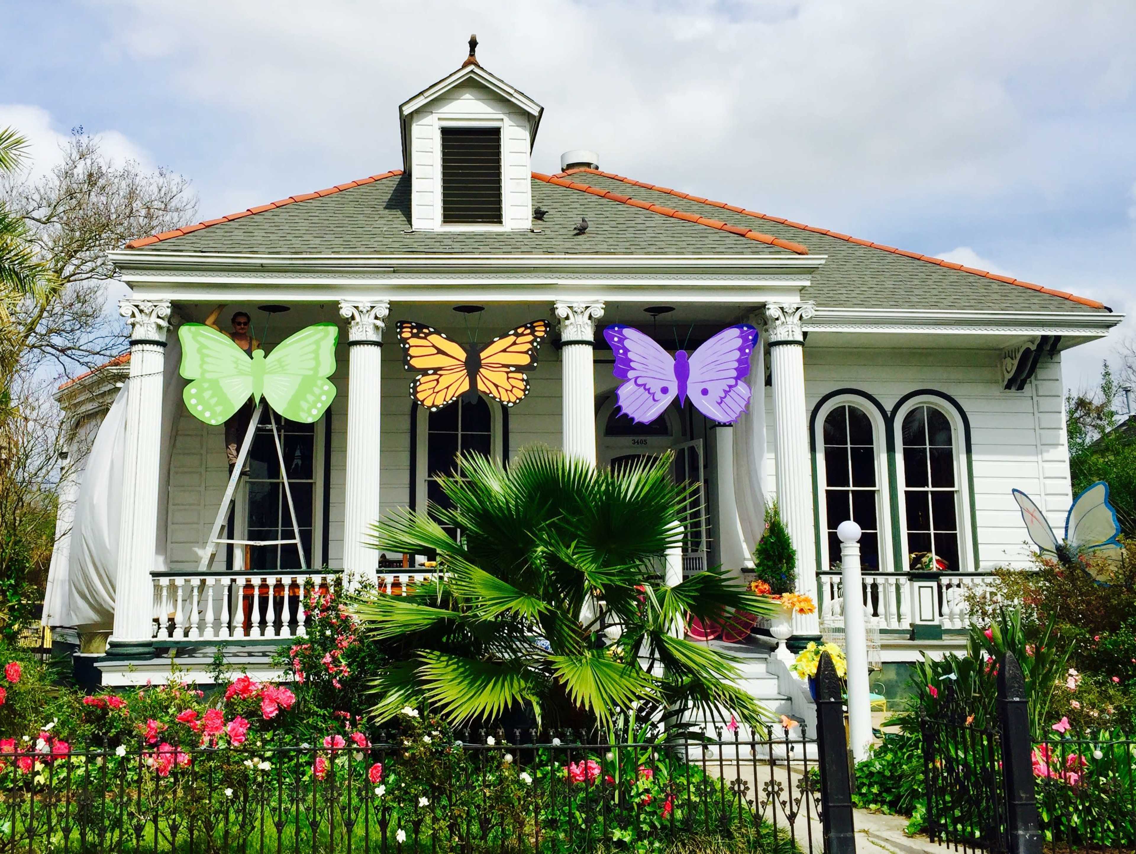 A white house with colorful butterfly decorations hangs from the porch, surrounded by a garden of flowers and palm trees.