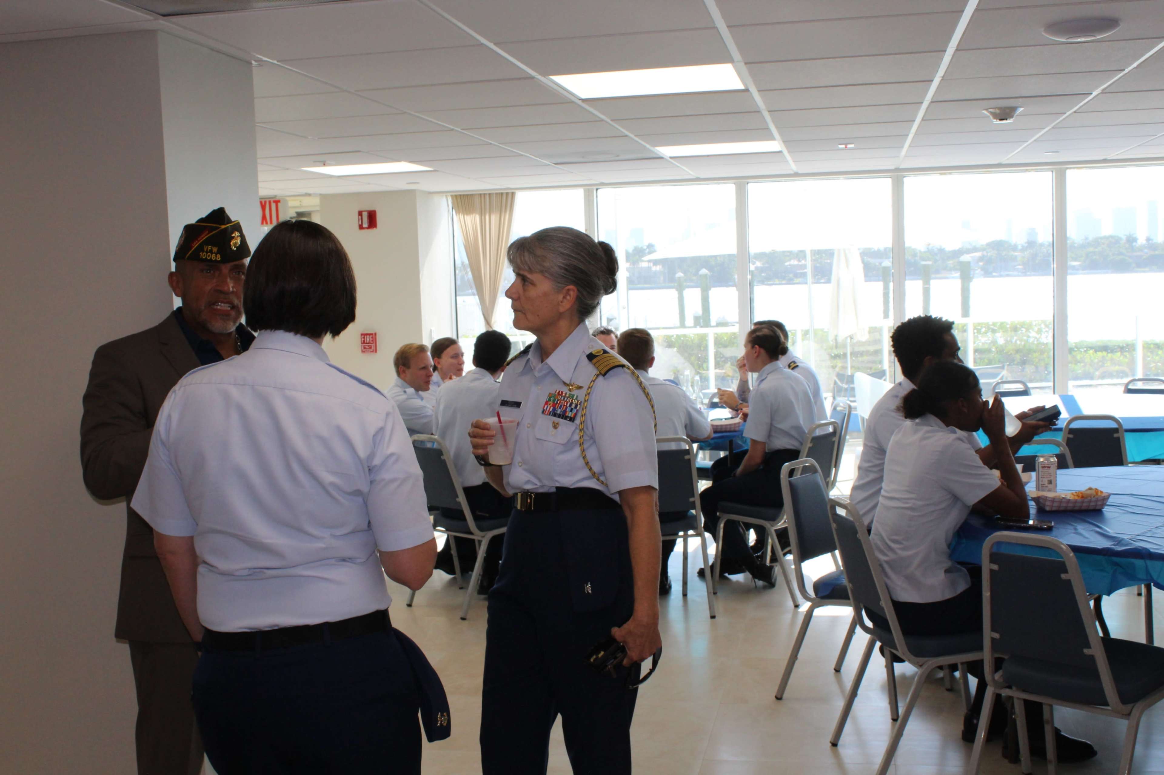 A group of military personnel and guests engage in conversation at a venue with tables set for an event and a view of a waterfront outside.