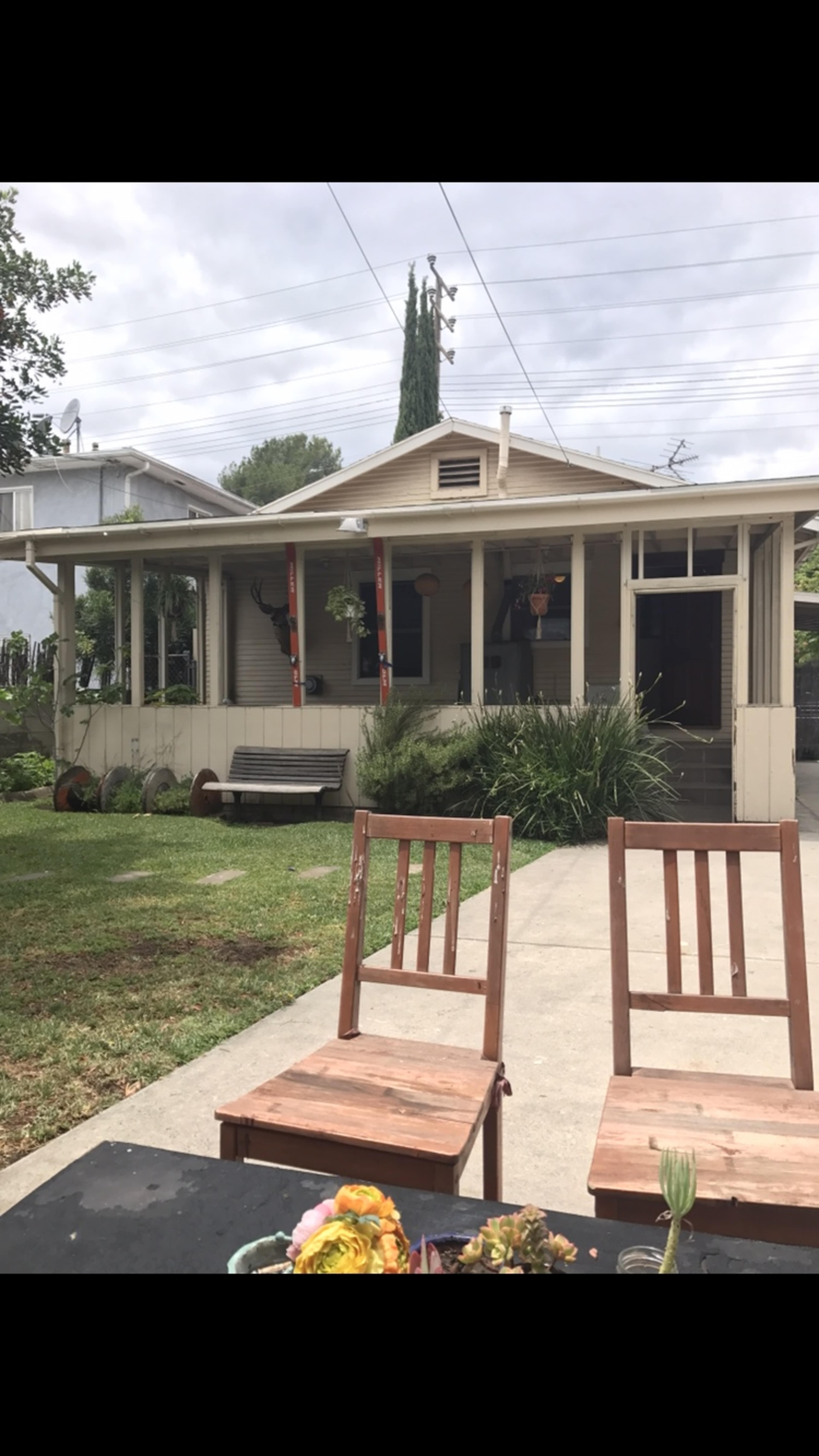 A wooden table with two empty chairs is positioned in front of a house featuring a porch and surrounding greenery.