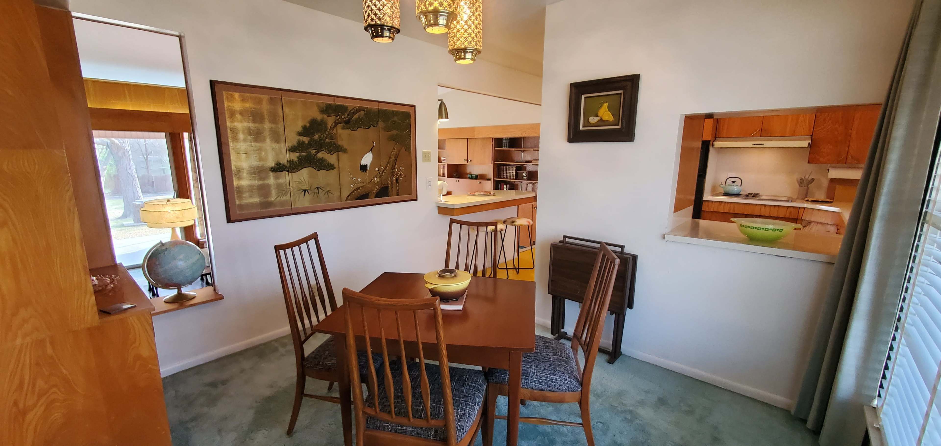 A dining area featuring a wooden table with six chairs, a painting on the wall, and a small kitchen space visible in the background.