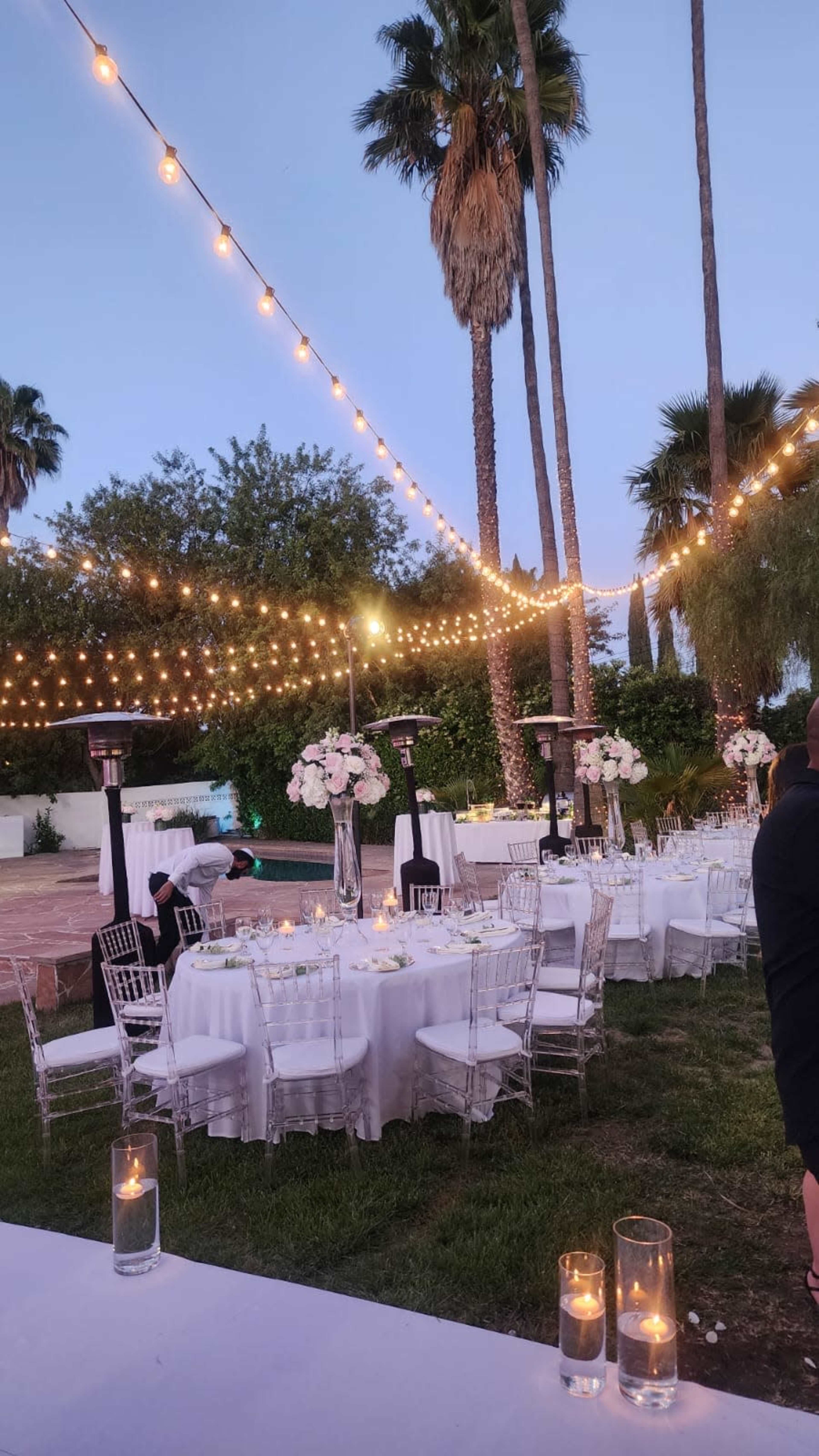 A beautifully arranged outdoor dining area featuring round tables set with white linens, surrounded by palm trees and illuminated by string lights.