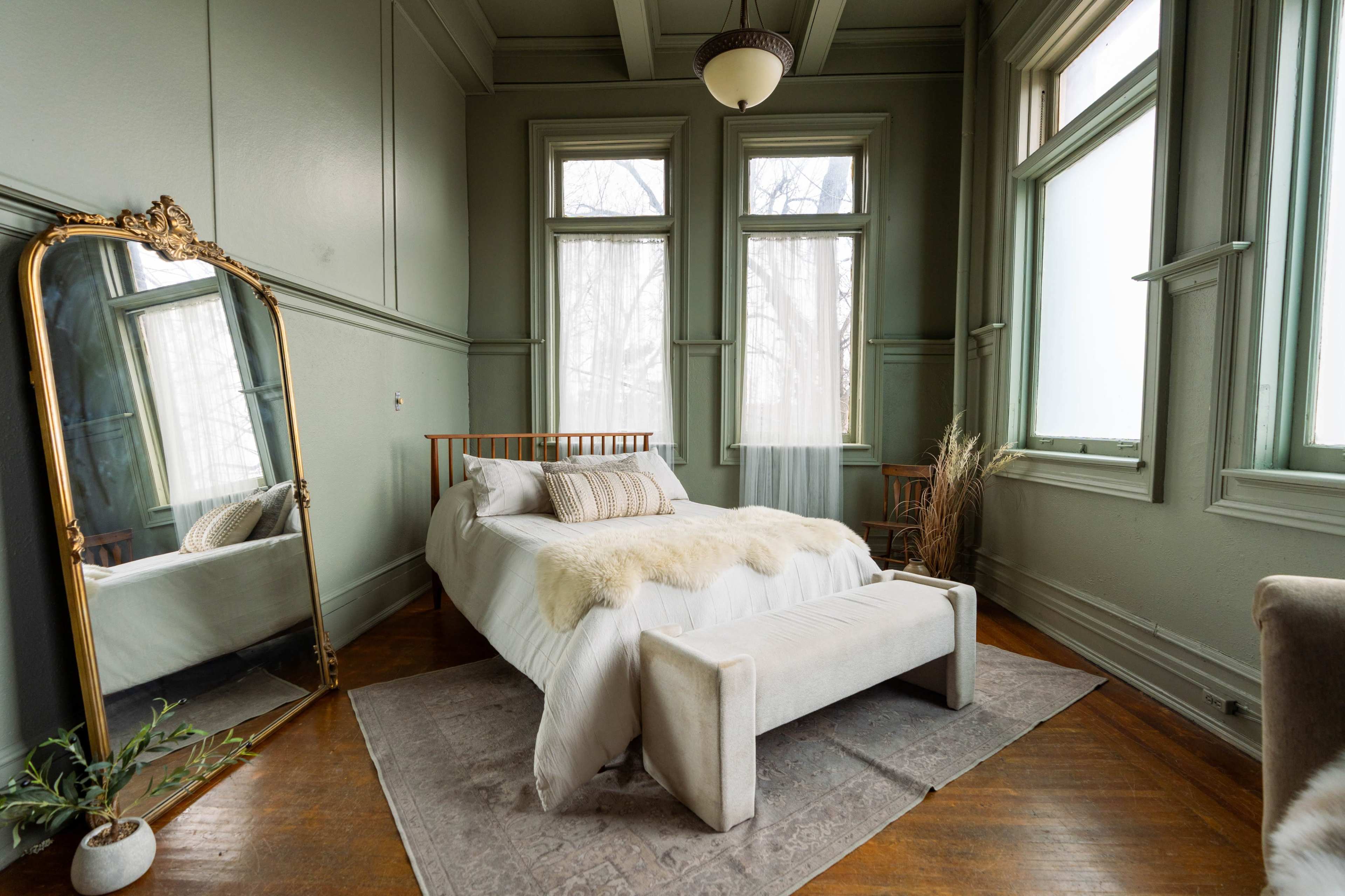 The room features a bed with a light-colored bedspread, large windows, a decorative mirror, and a cozy armchair.
