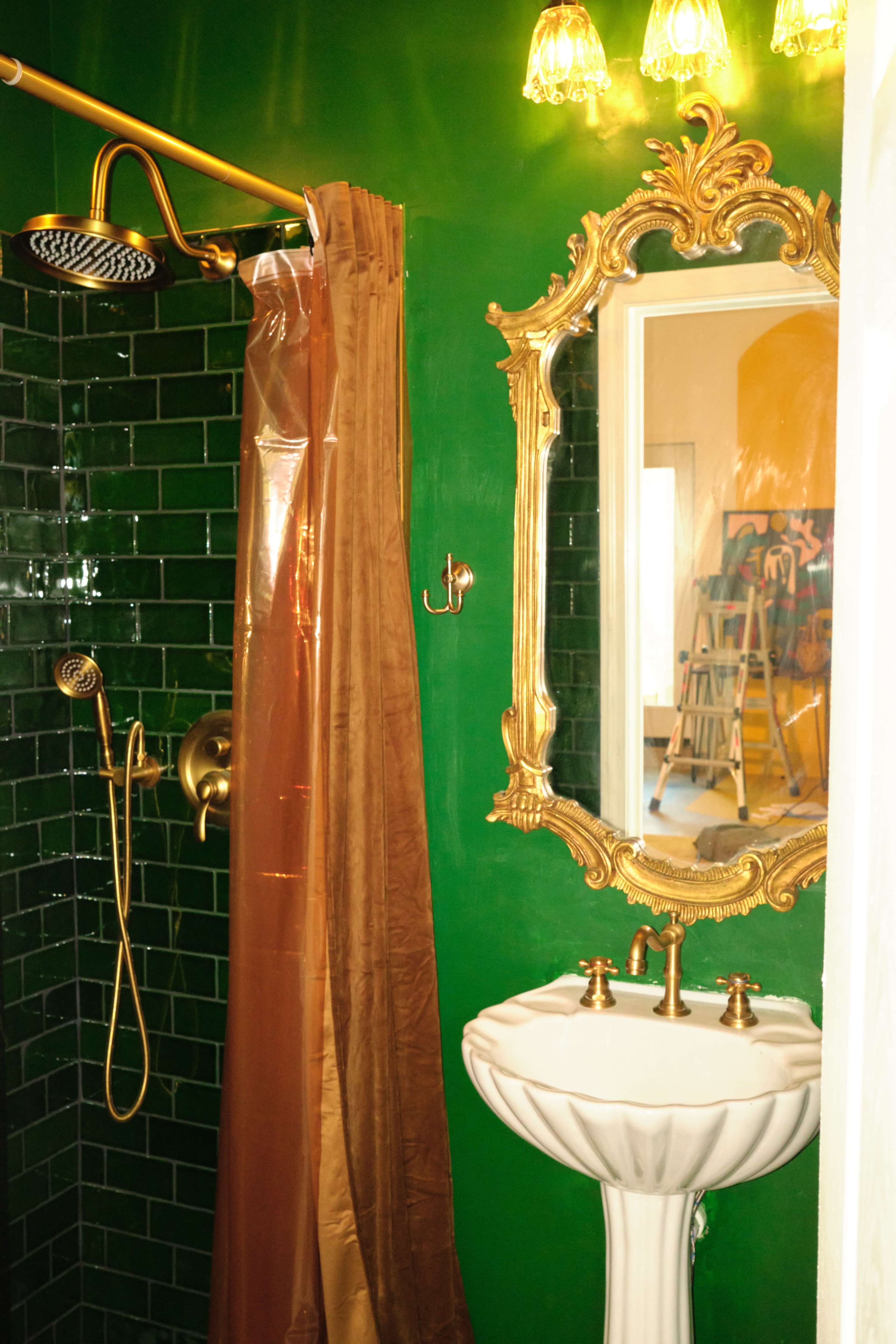 The image shows a bathroom with green tiled walls, a golden shower fixture, a decorative mirror, and a white pedestal sink.