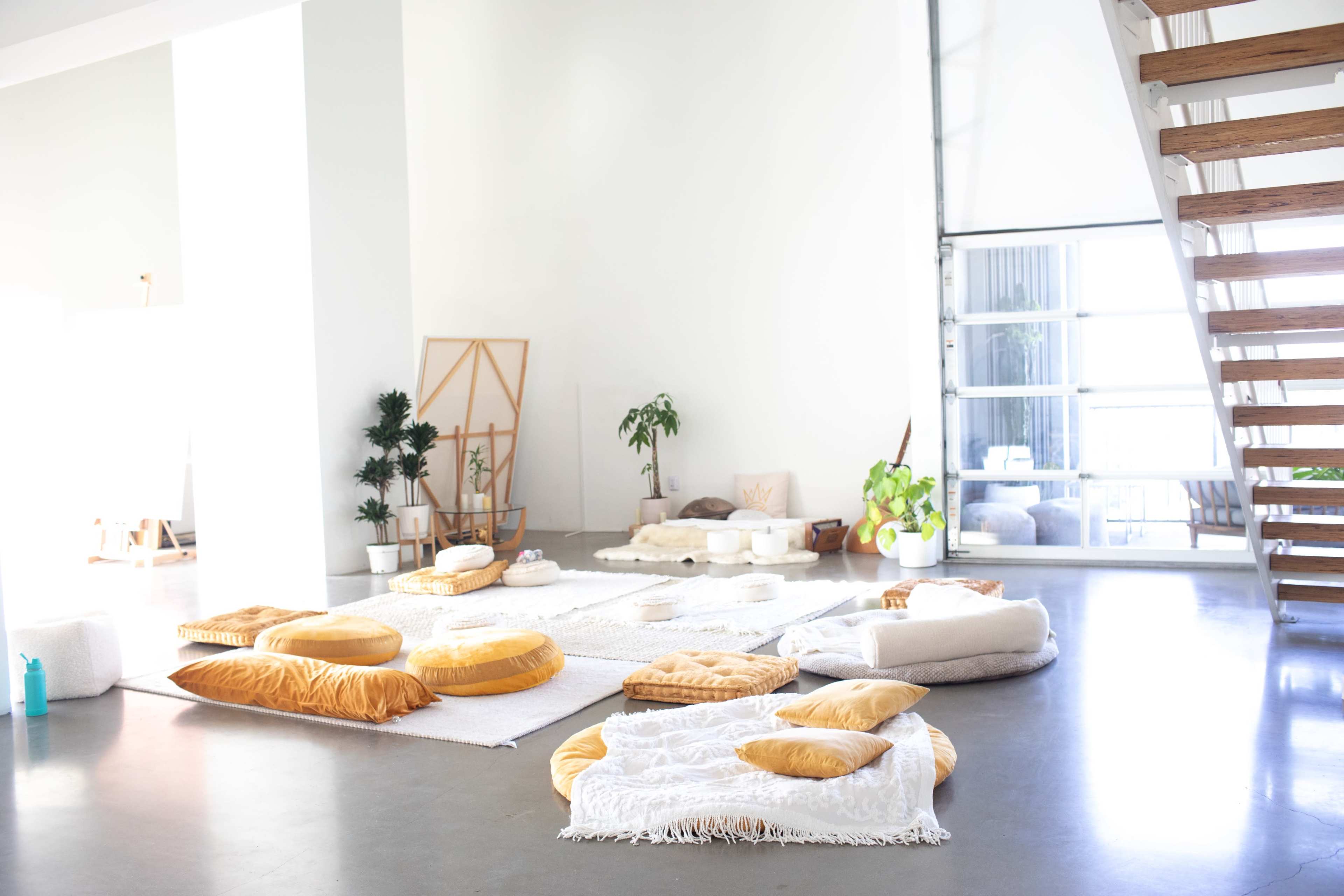 A spacious, sunlit room with various floor cushions arranged on a white rug, accompanied by plants and a staircase.