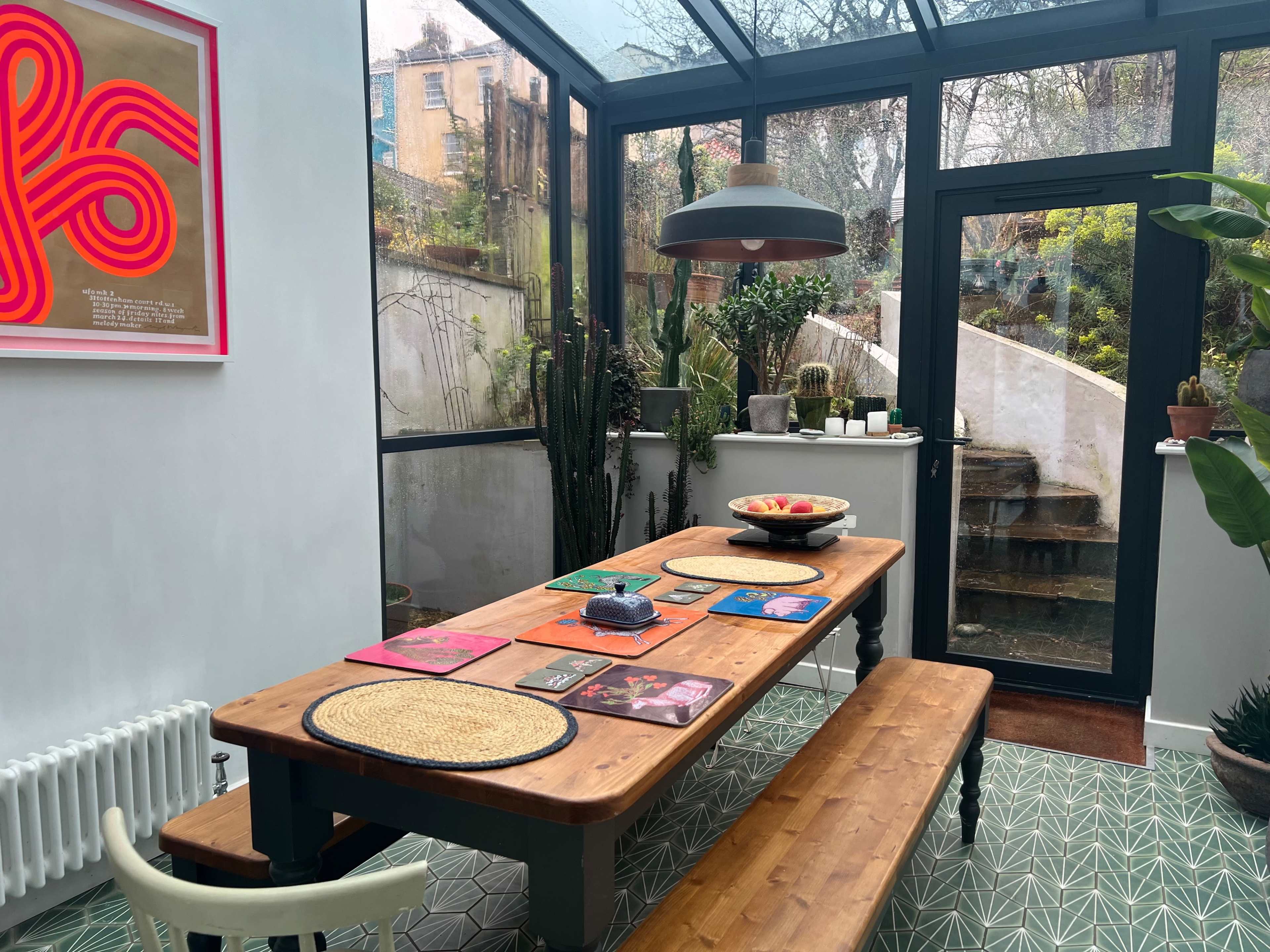 A wooden dining table with benches is set in a bright room featuring large glass windows and a view of a garden, adorned with plants and artwork on the walls.
