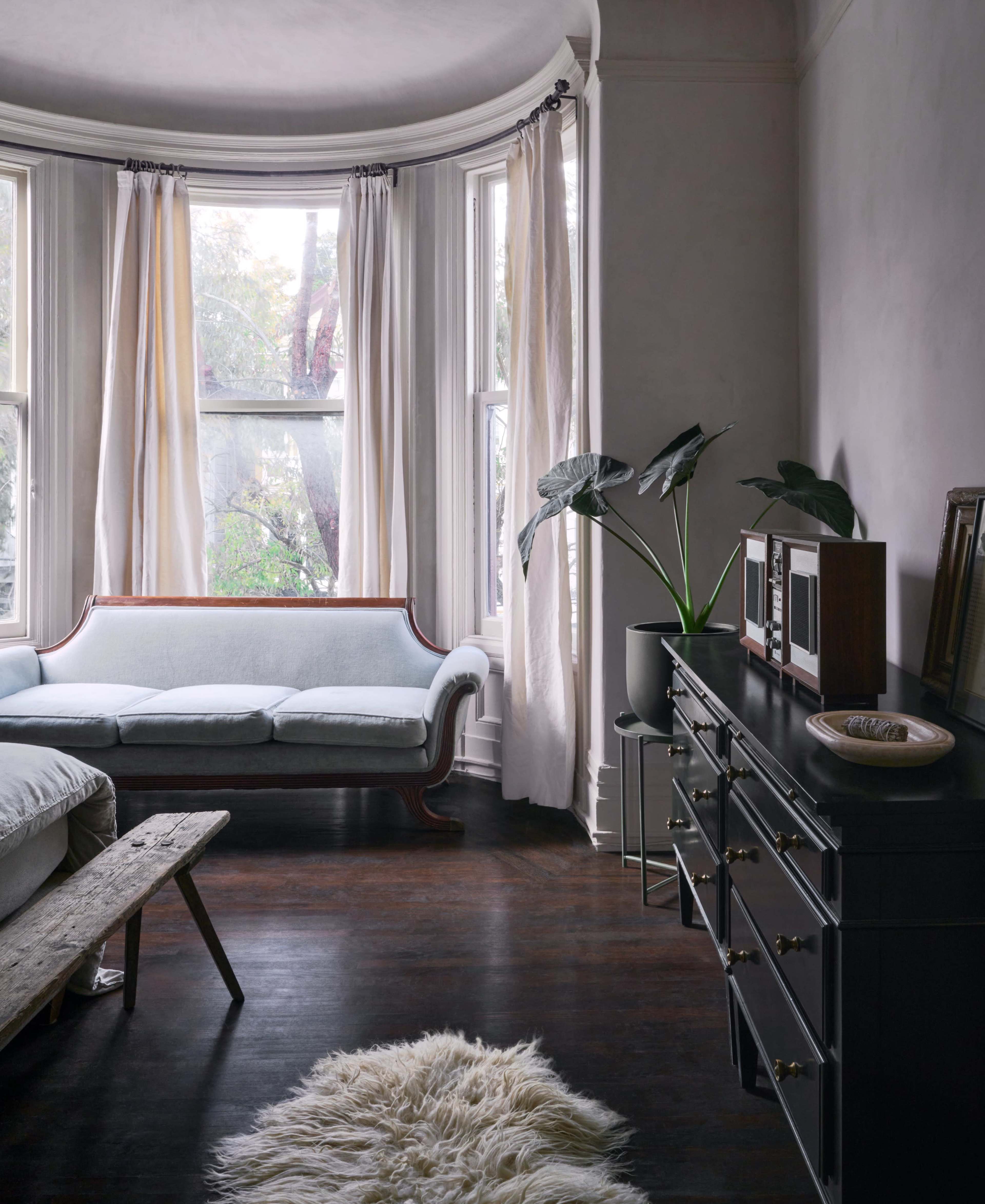 A light-filled living room features a vintage sofa by a large window, a wooden bench, a dark dresser, and a potted plant.