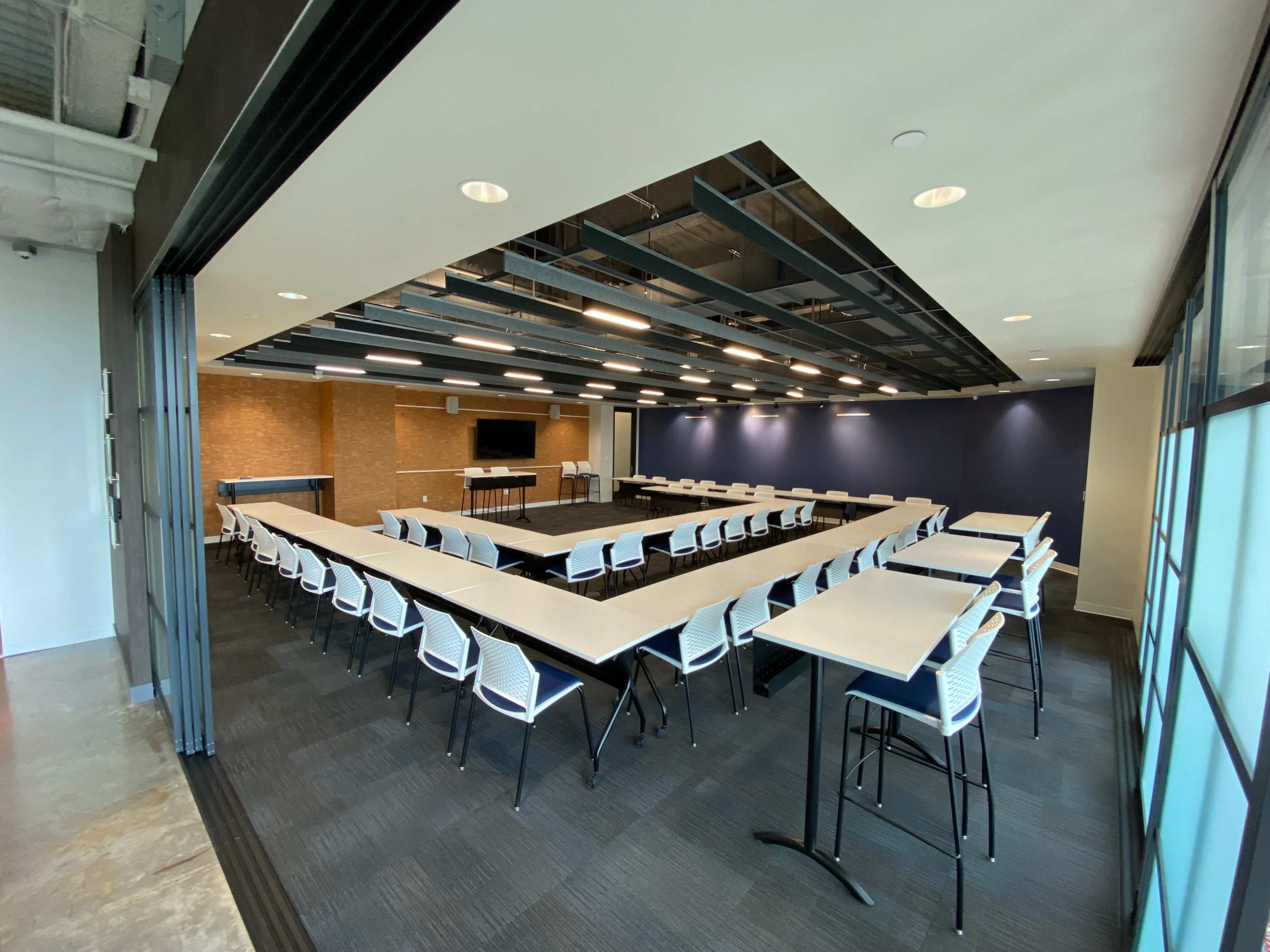 A modern conference room arranged in a U-shape with white chairs and tables, featuring a large screen at one end and dark blue walls.