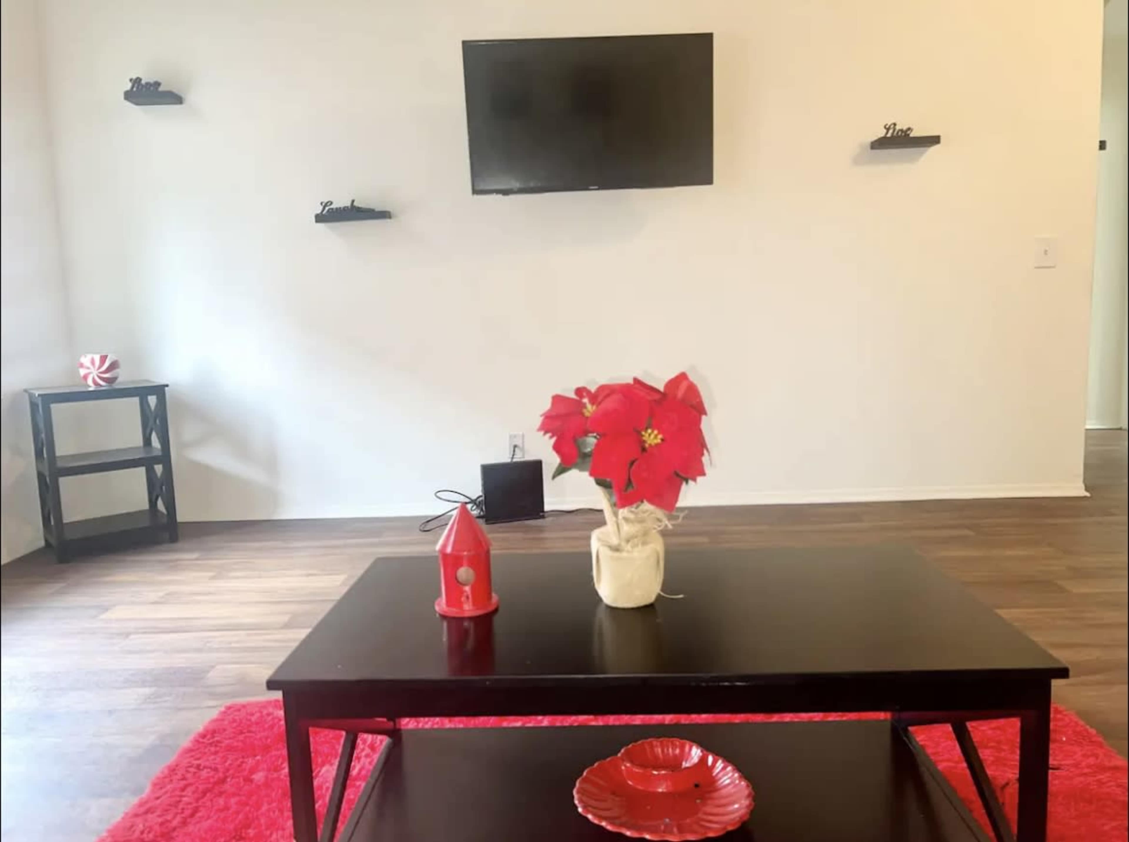 The scene depicts a living room with a black coffee table, a red rug, a television mounted on the wall, and decorative items, including a plant and a red candle holder.