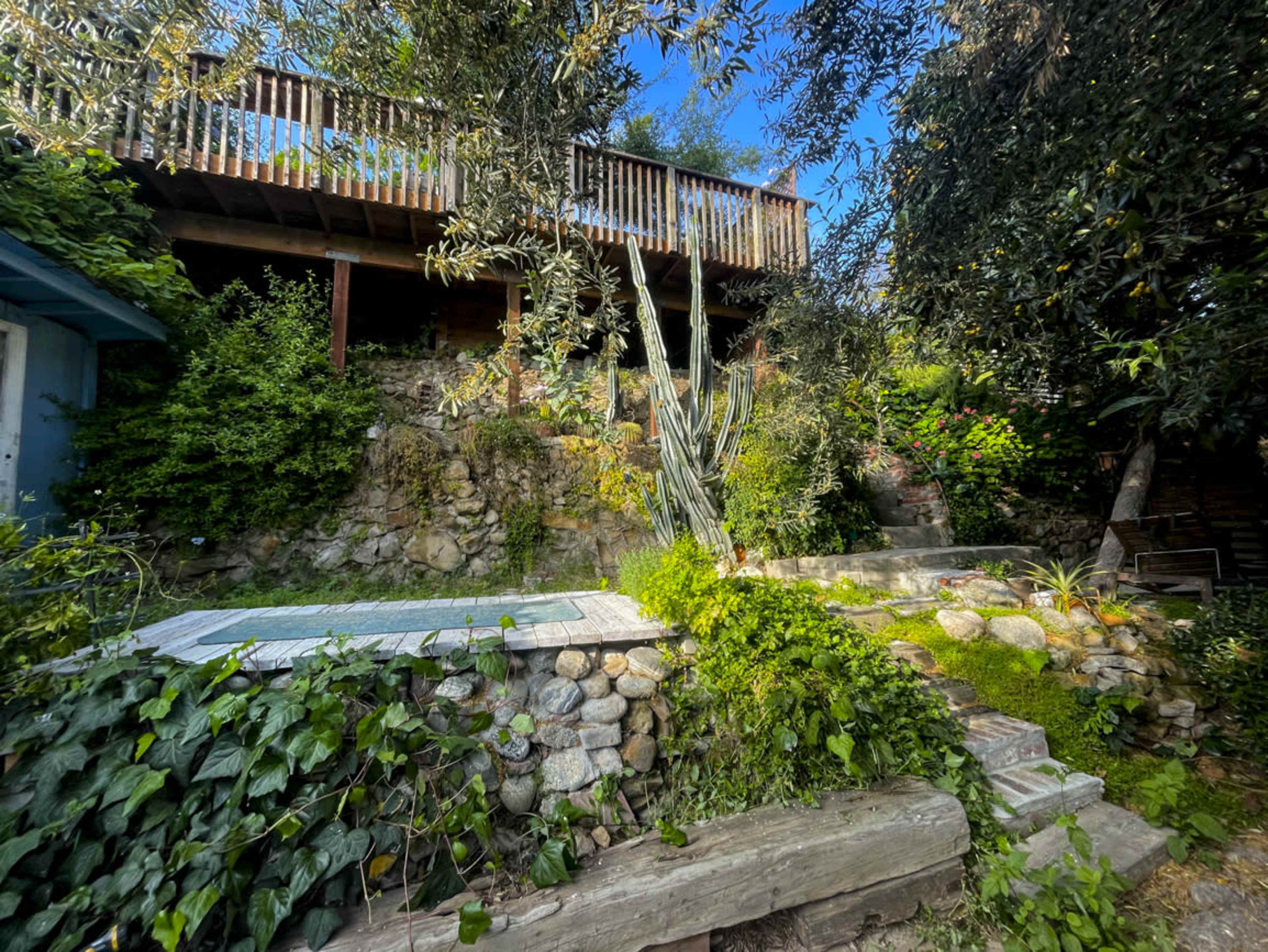 The image shows a stone-walled garden with a small swimming pool, surrounded by various plants and a staircase leading to a deck above.