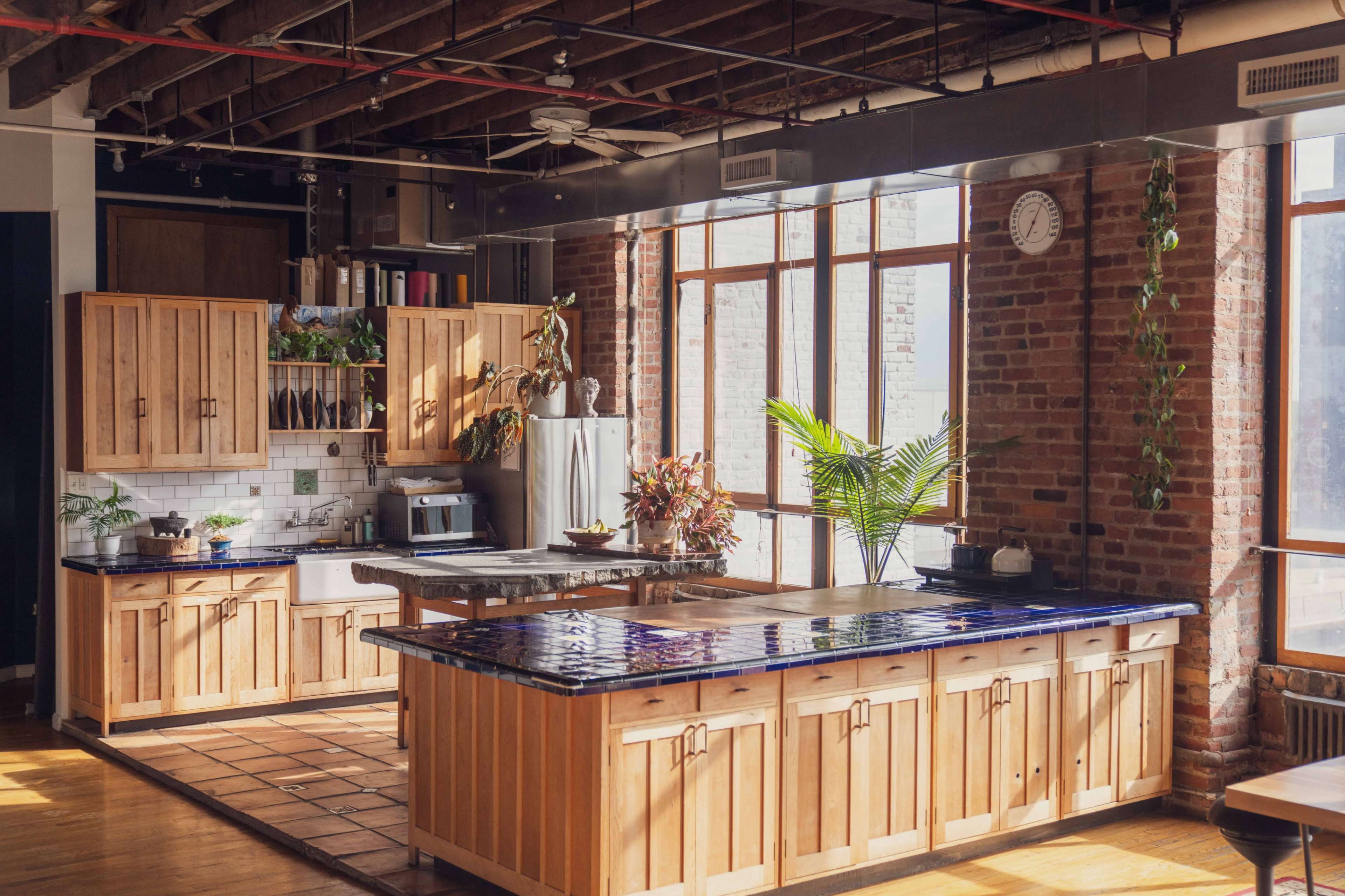 The kitchen features wooden cabinets, a tiled island, and large windows that bring in natural light, all set against exposed brick walls.
